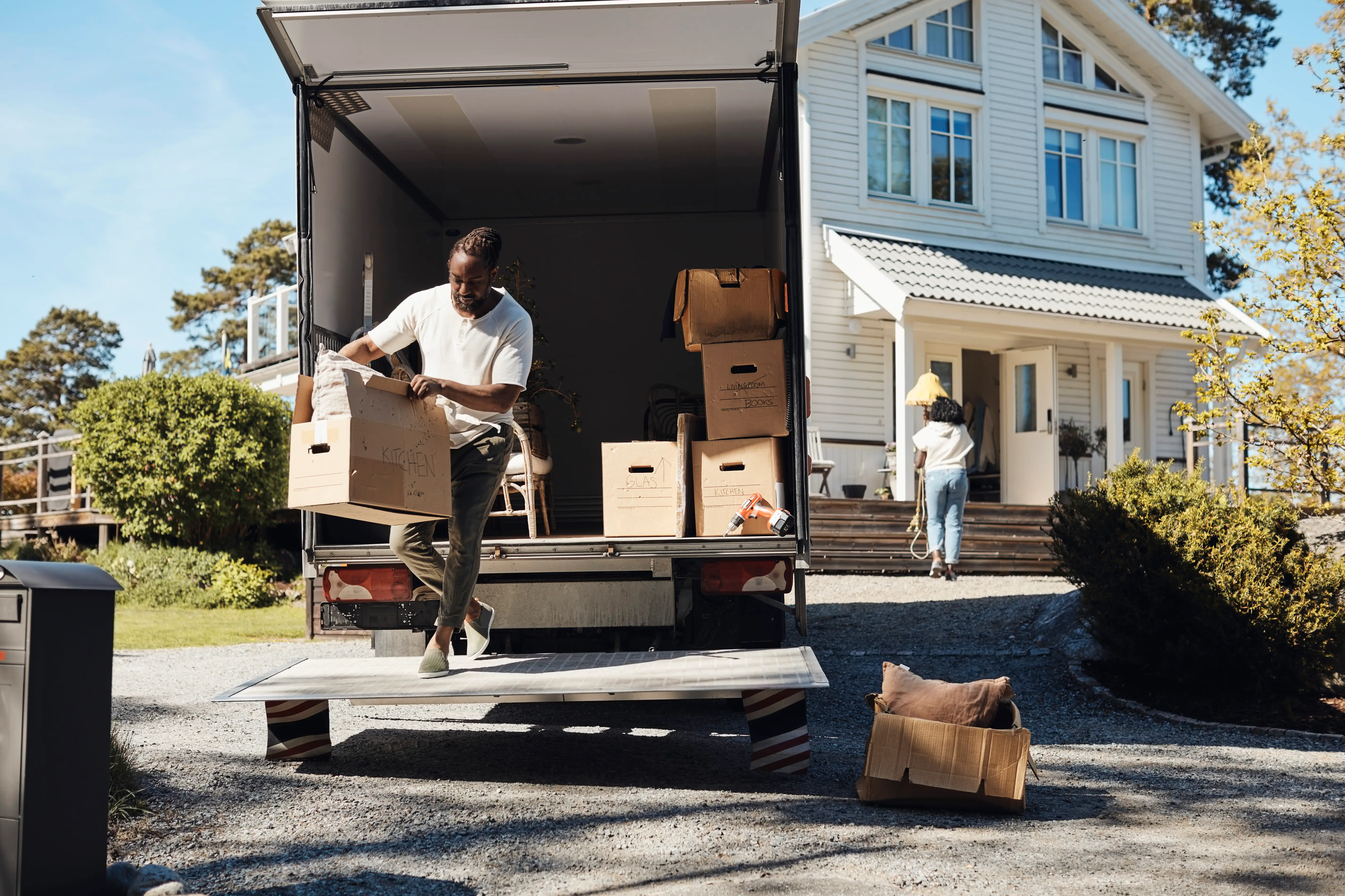A man moves boxes out of a moving truck. In the background, a woman walks toward the front door of a large house.