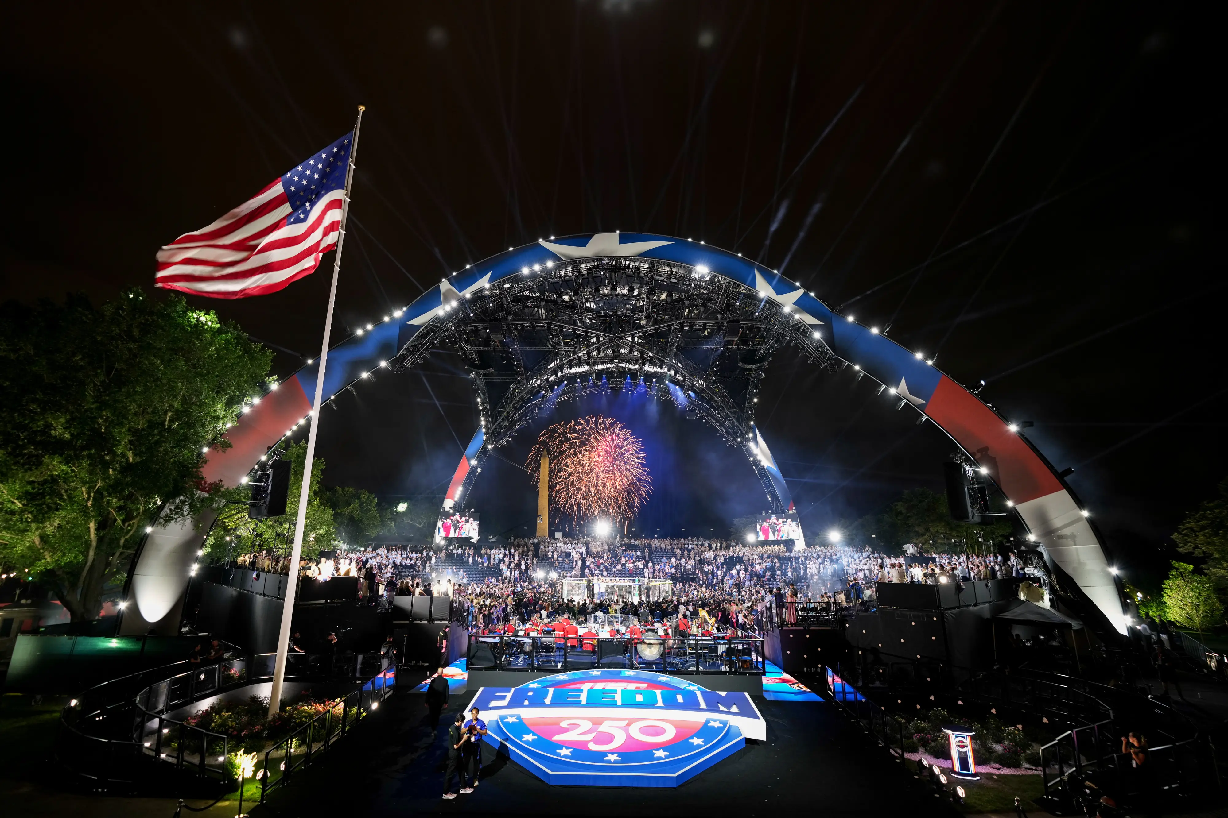 Fireworks during the UFC fight on the White House lawn with a flag flying in the foreground and a Freedom 250 sign.