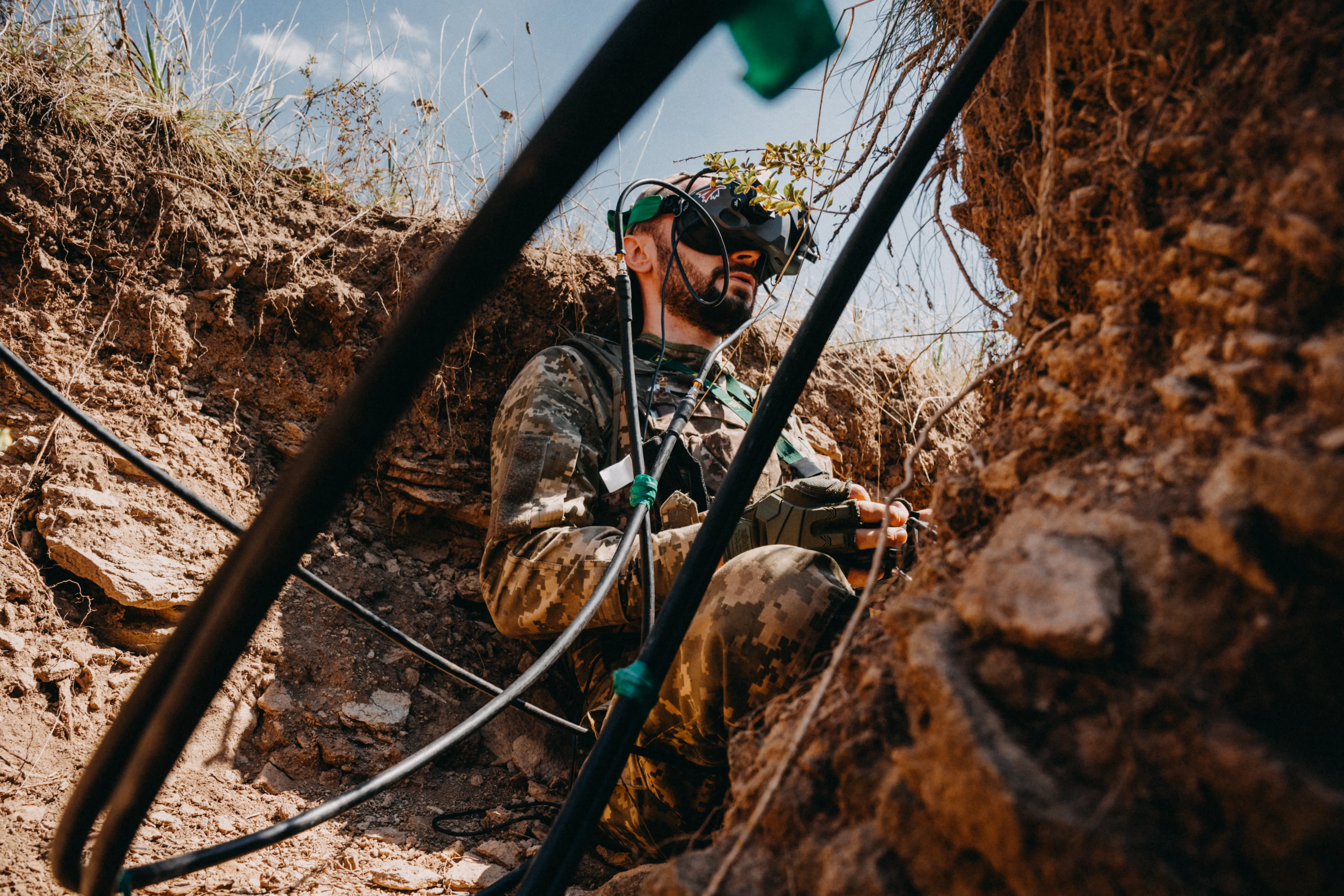 A man in camouflage gear and wearing a headset sits in a trench with a blue sky above him