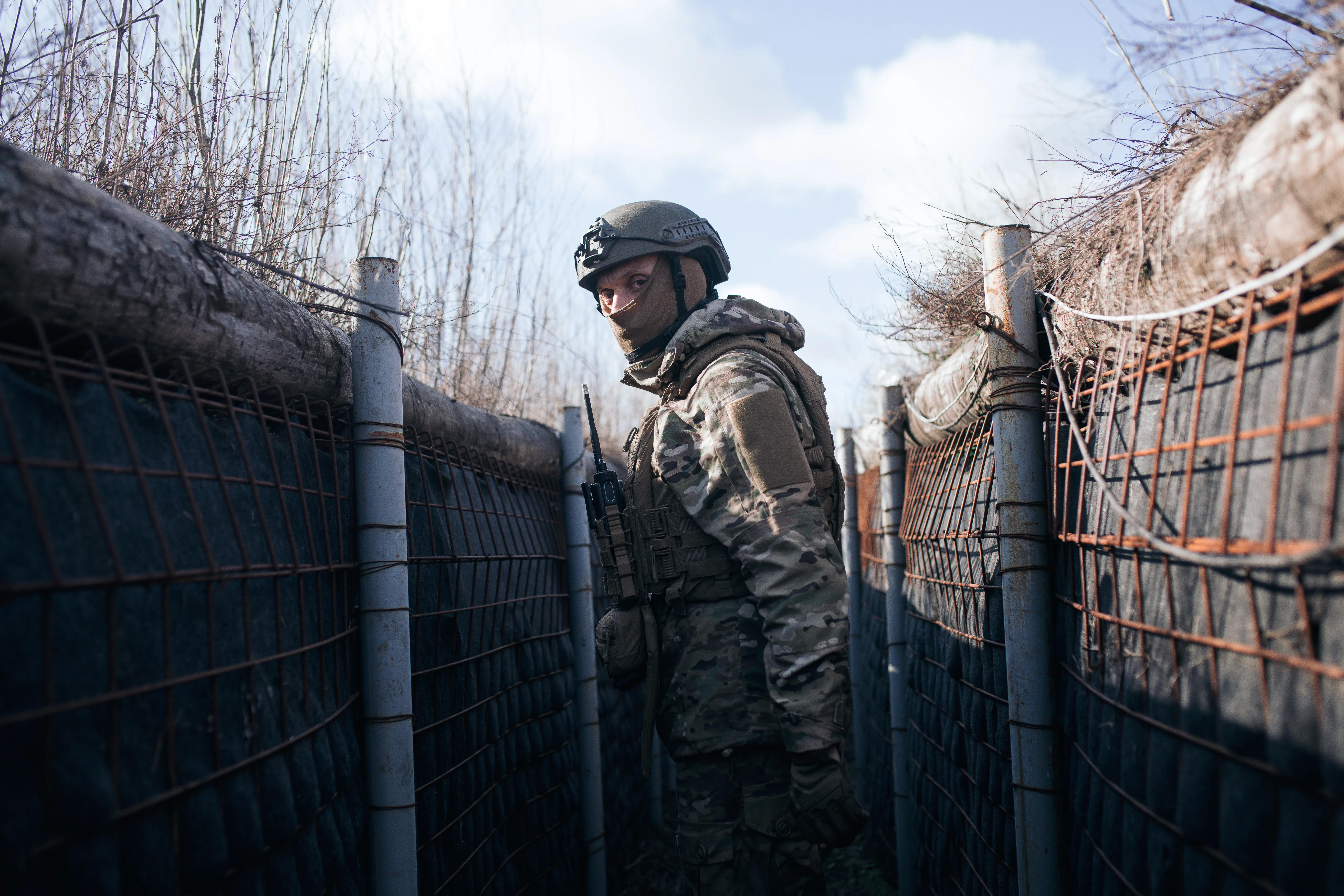 A man in camouflage and a helmet stands in a trench under a blue sky