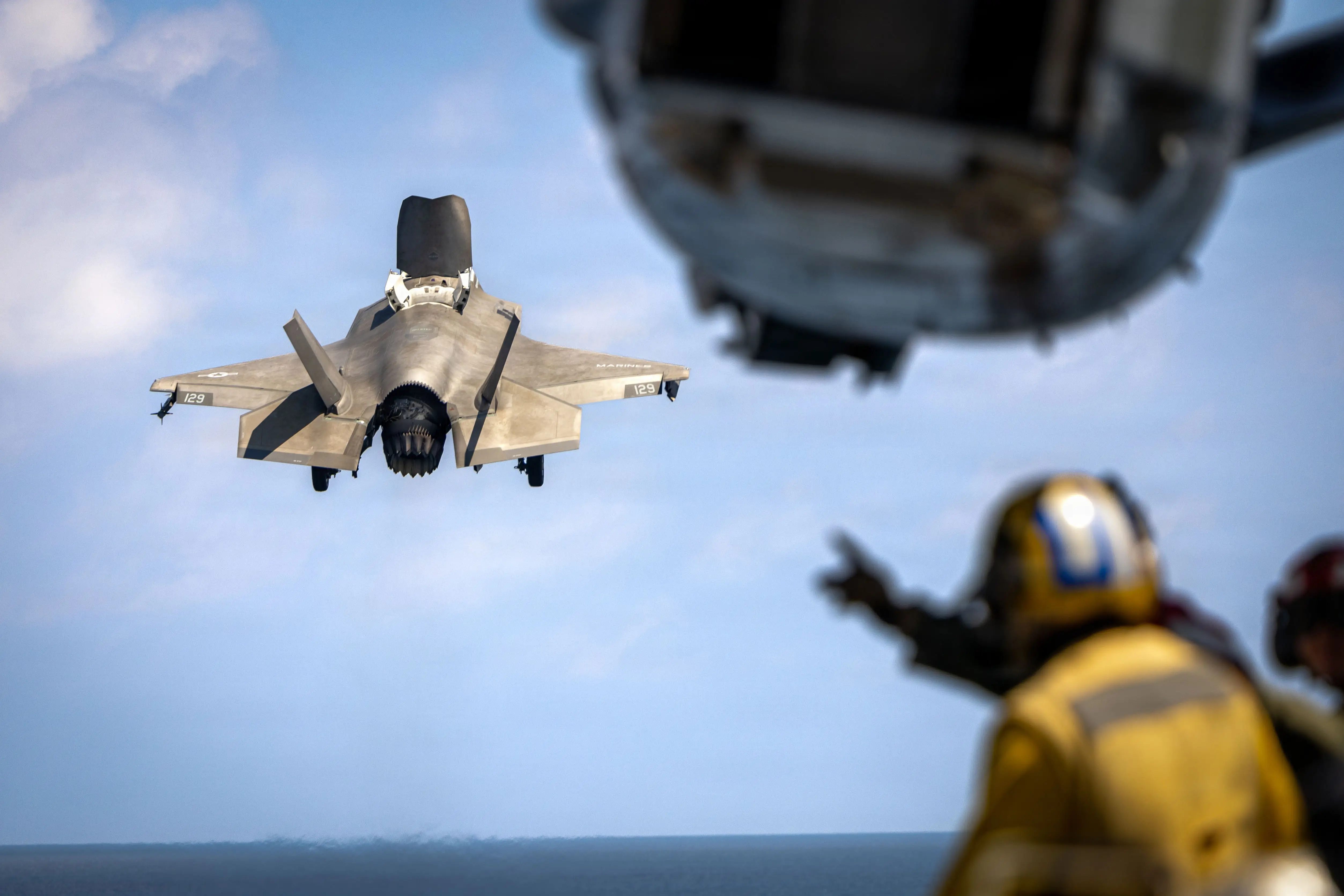 A US F-35 aircraft flies in a blue cloudy sky. A man points at it from the deck of a ship.