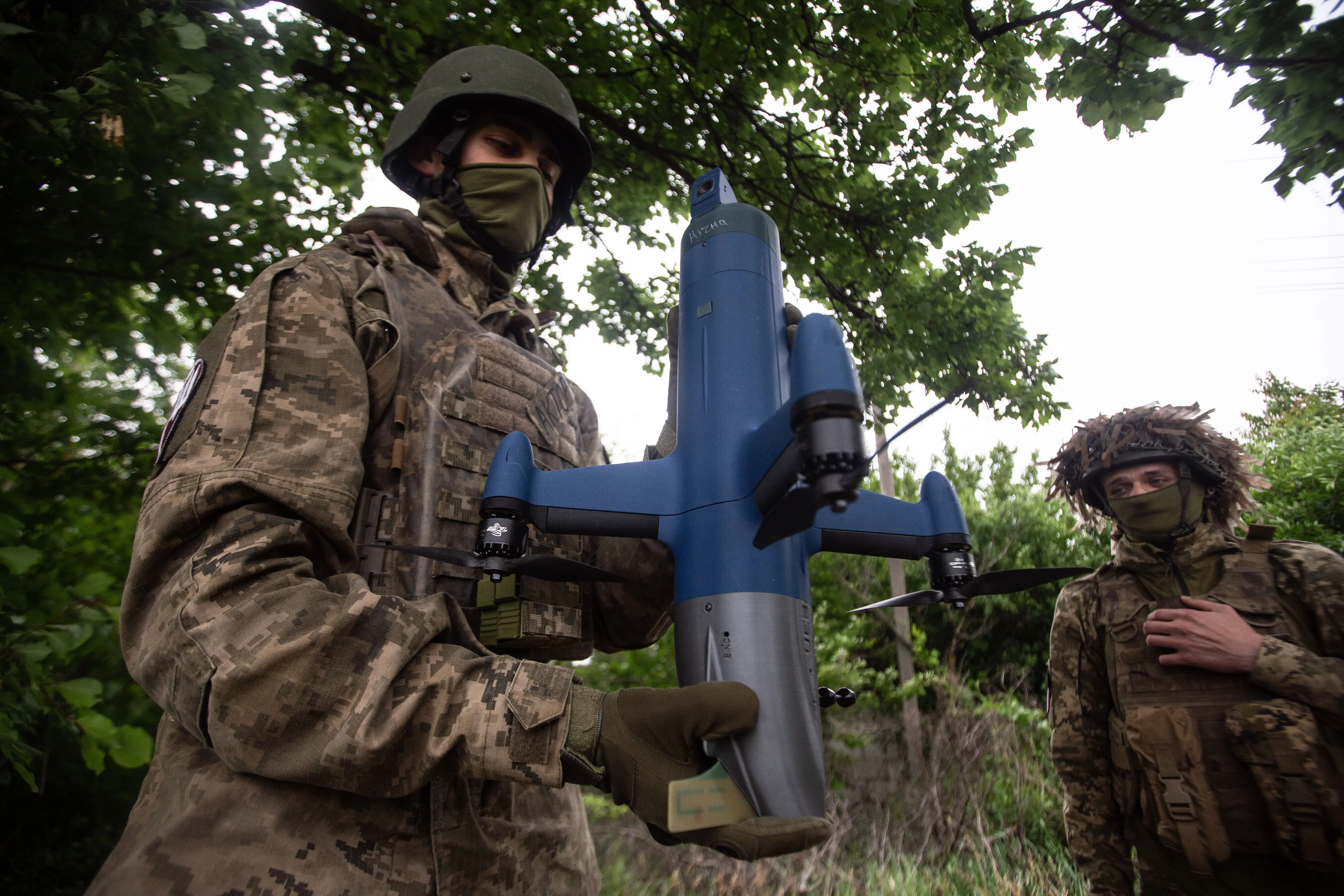 Two men in camouflage gear, helmets, and face scarves stand under trees, with one holding a blue interceptor drone