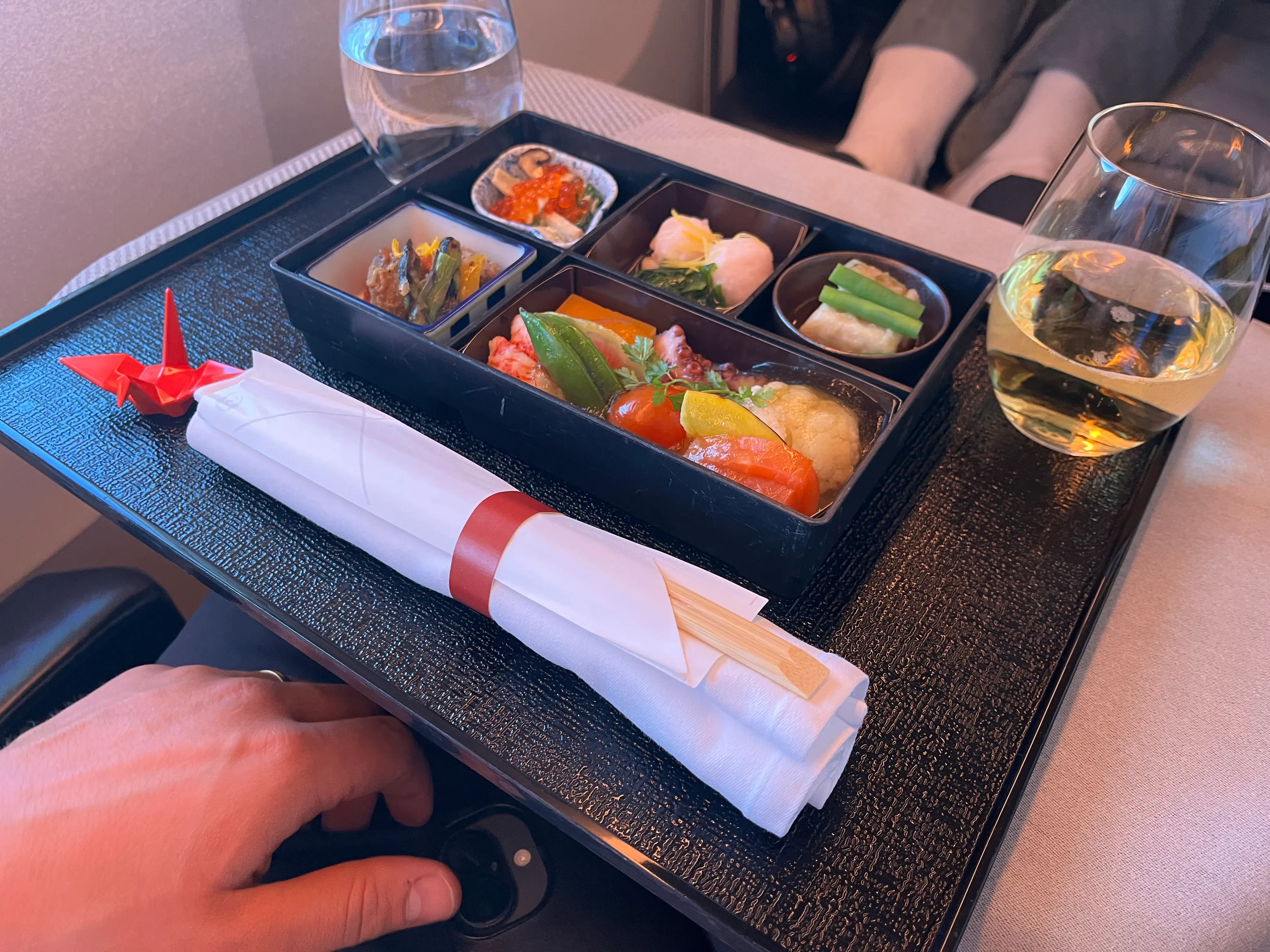Tray on plane with wine, wter, origami swan, and bento box of food with chopsticks, napkin