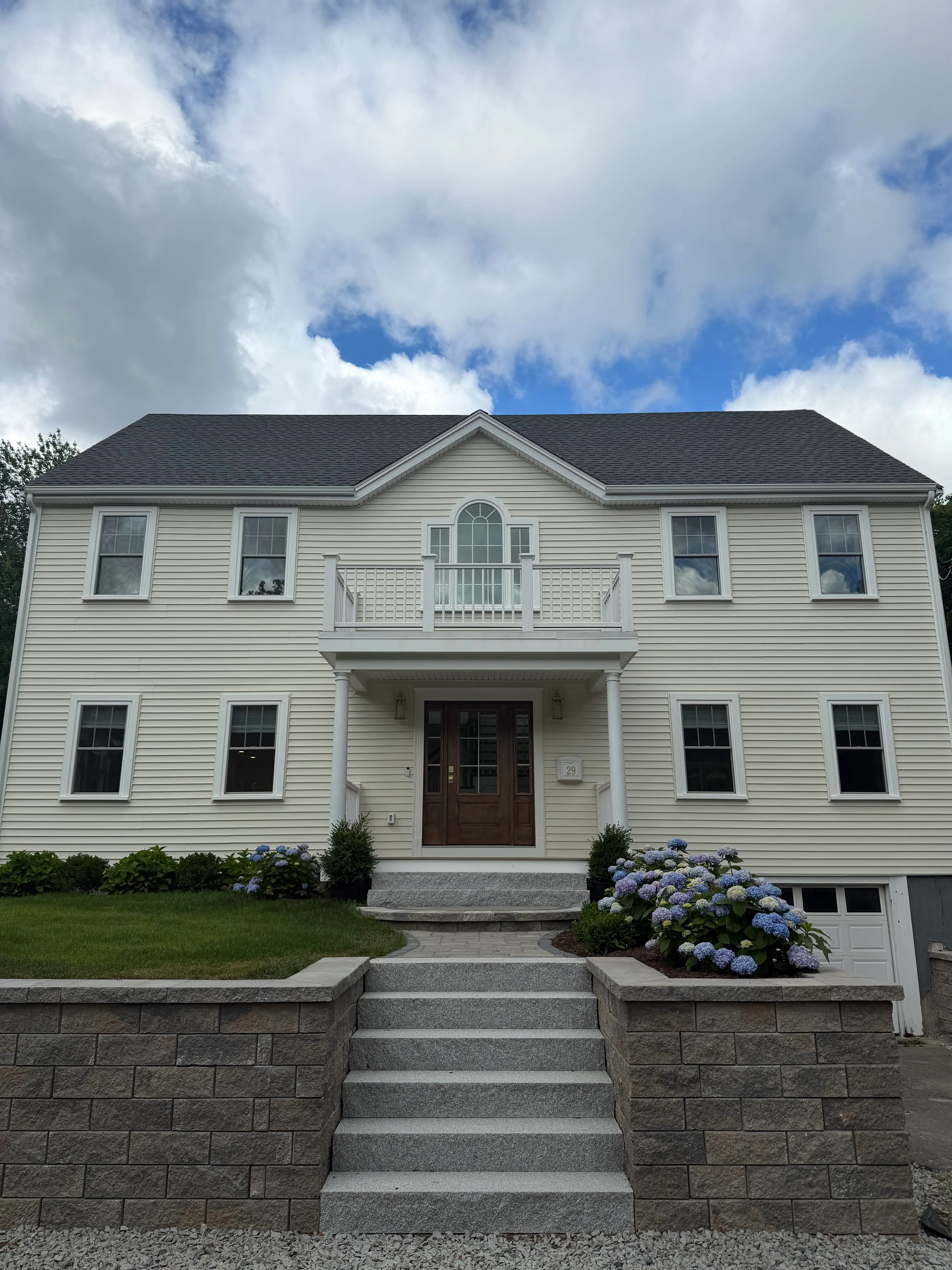 A two-story colonial home with a staircase leading to it and hydrangeas in the front lawn.