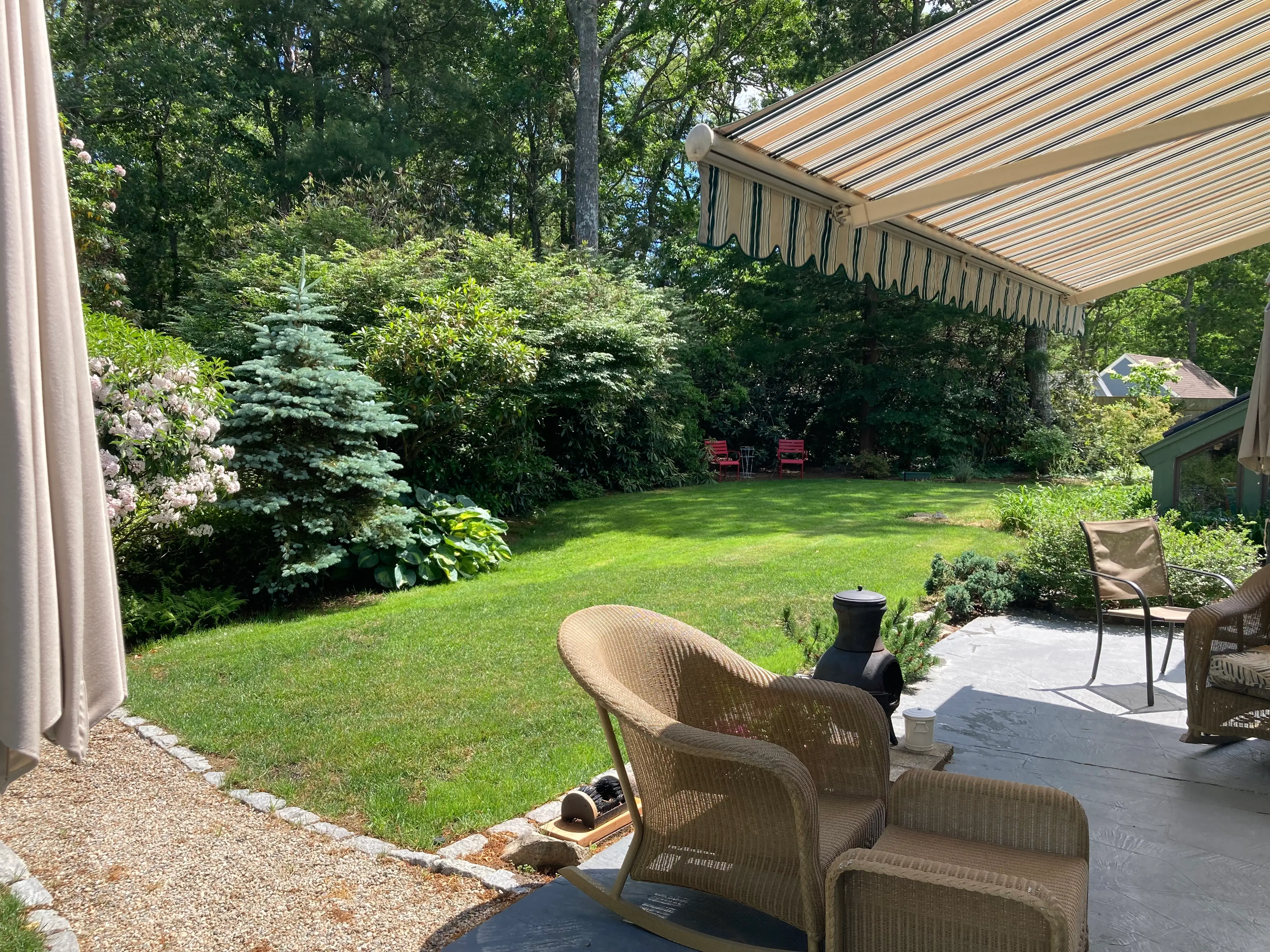 Shaded patio with wicker seating overlooks a green backyard lawn surrounded by dense trees and garden shrubs.