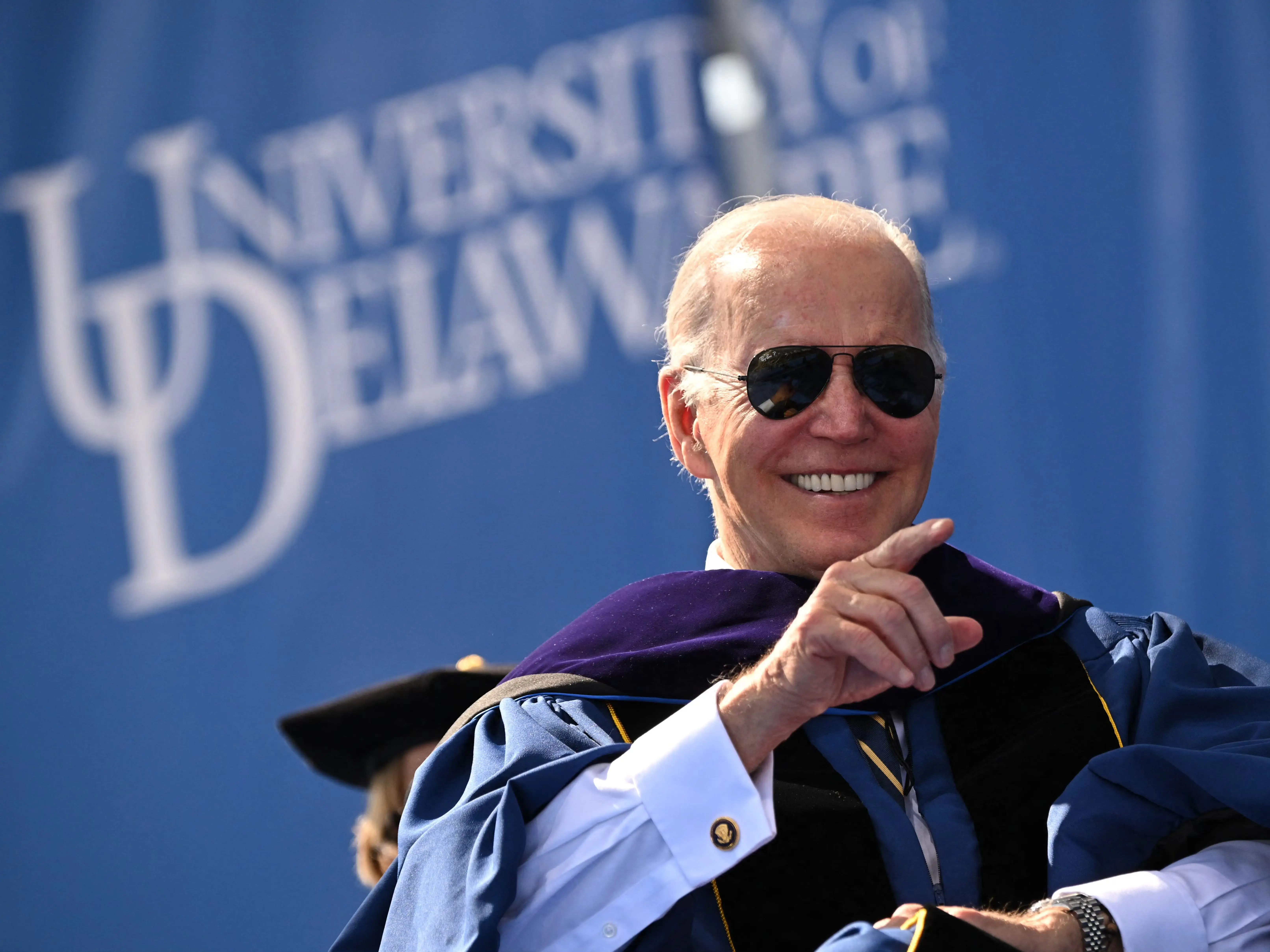Joe Biden smiles in sunglasses and academic regalia before a University of Delaware backdrop.