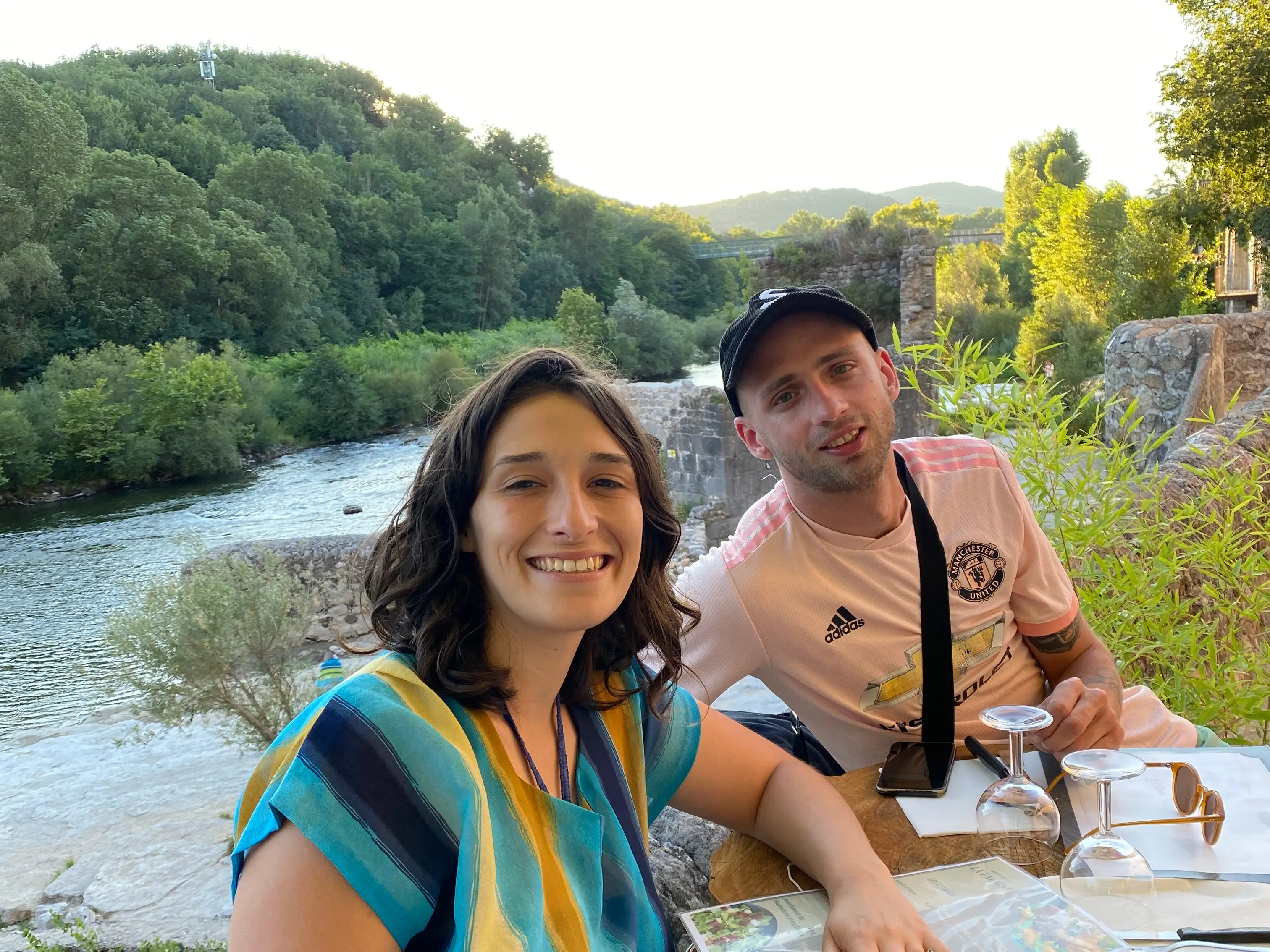The writer and her husband sitting at a table with wineglasses, with a body of water and trees in the background.