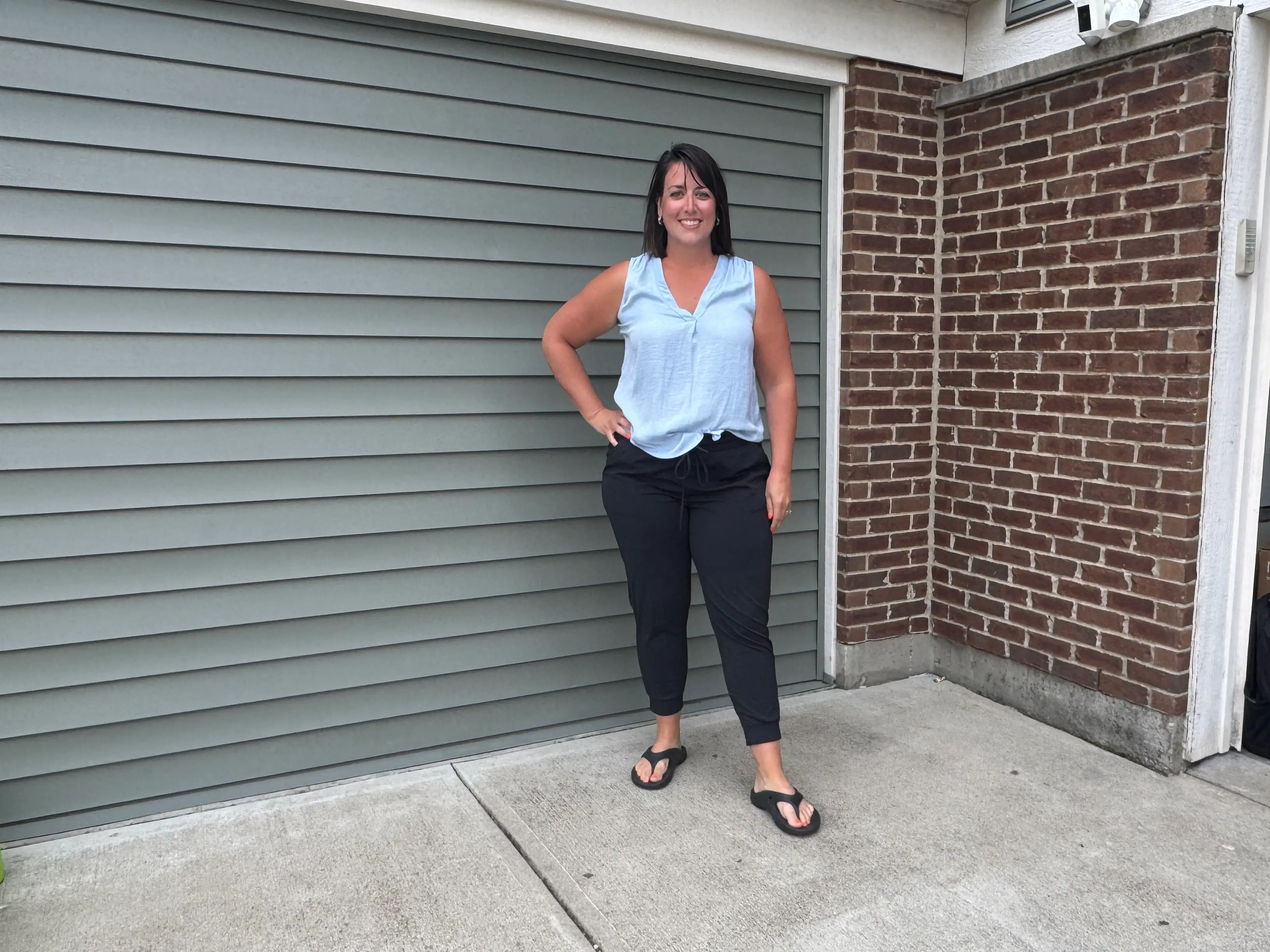 Woman in blue shirt, black pants posing in front of garage and brick wall