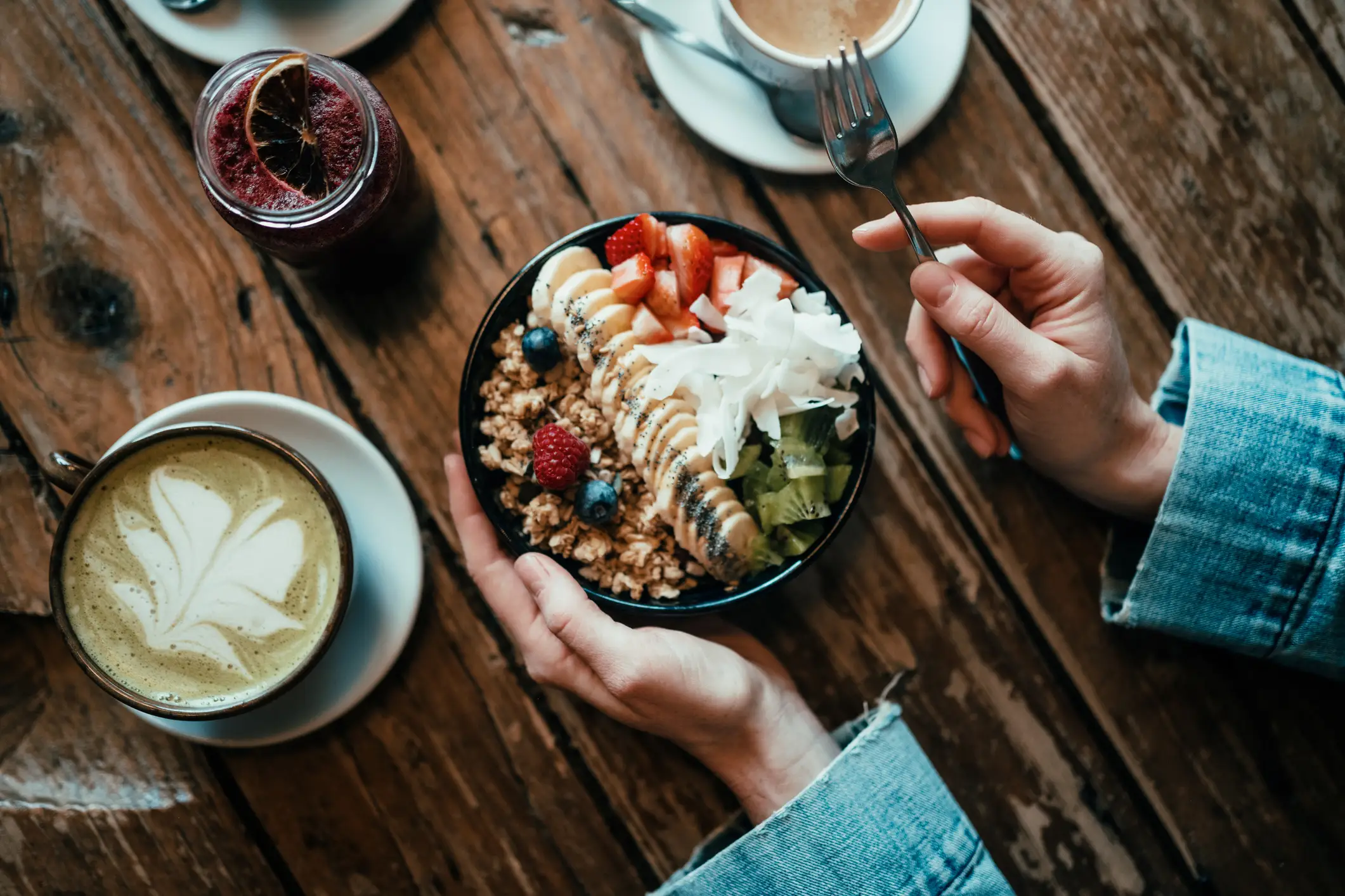 Hands hold a fruit and granola bowl beside coffee cups and a red drink on a rustic wooden table.