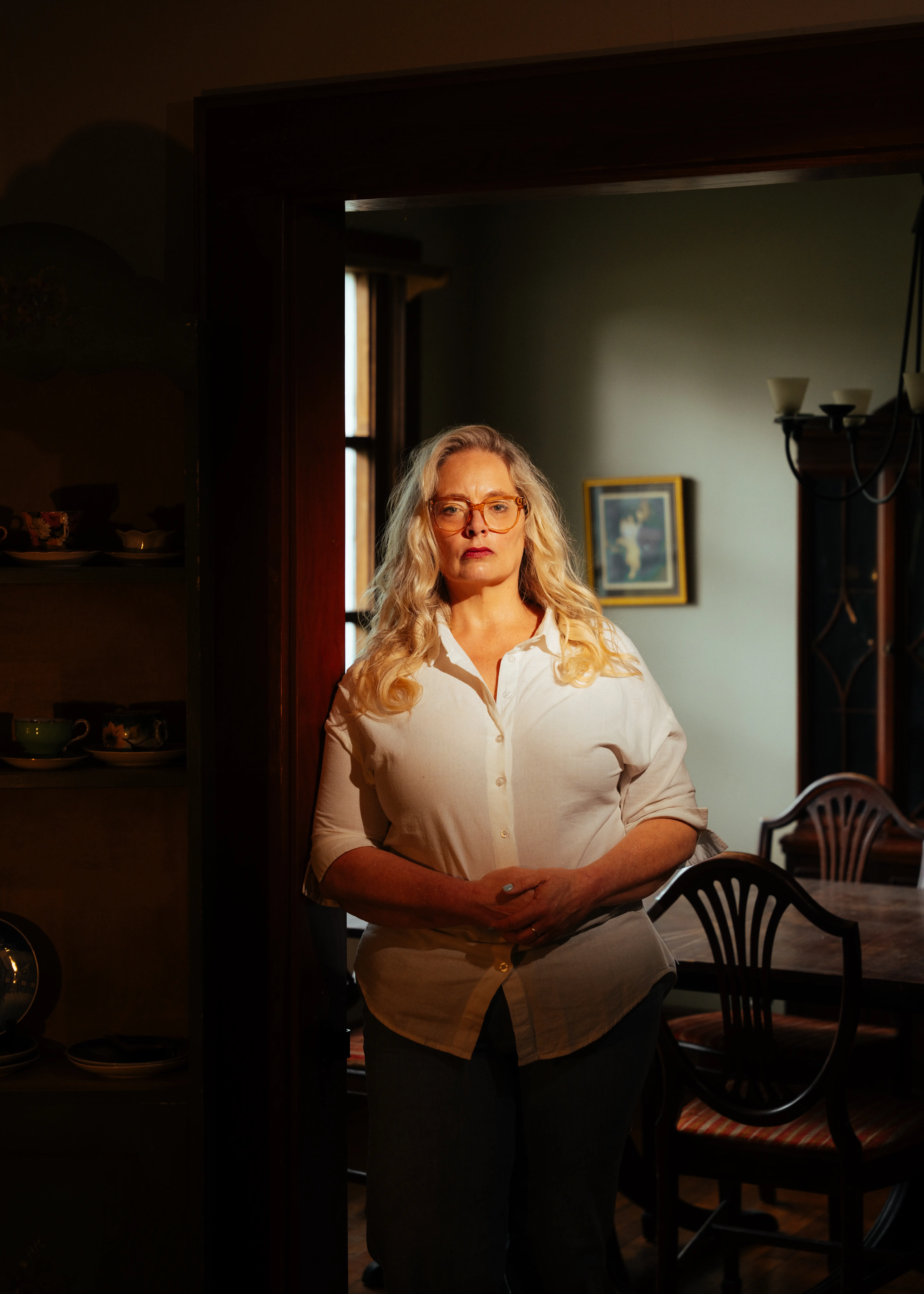Person with glasses stands in warm sunlight in a doorway beside a dining table and chairs.