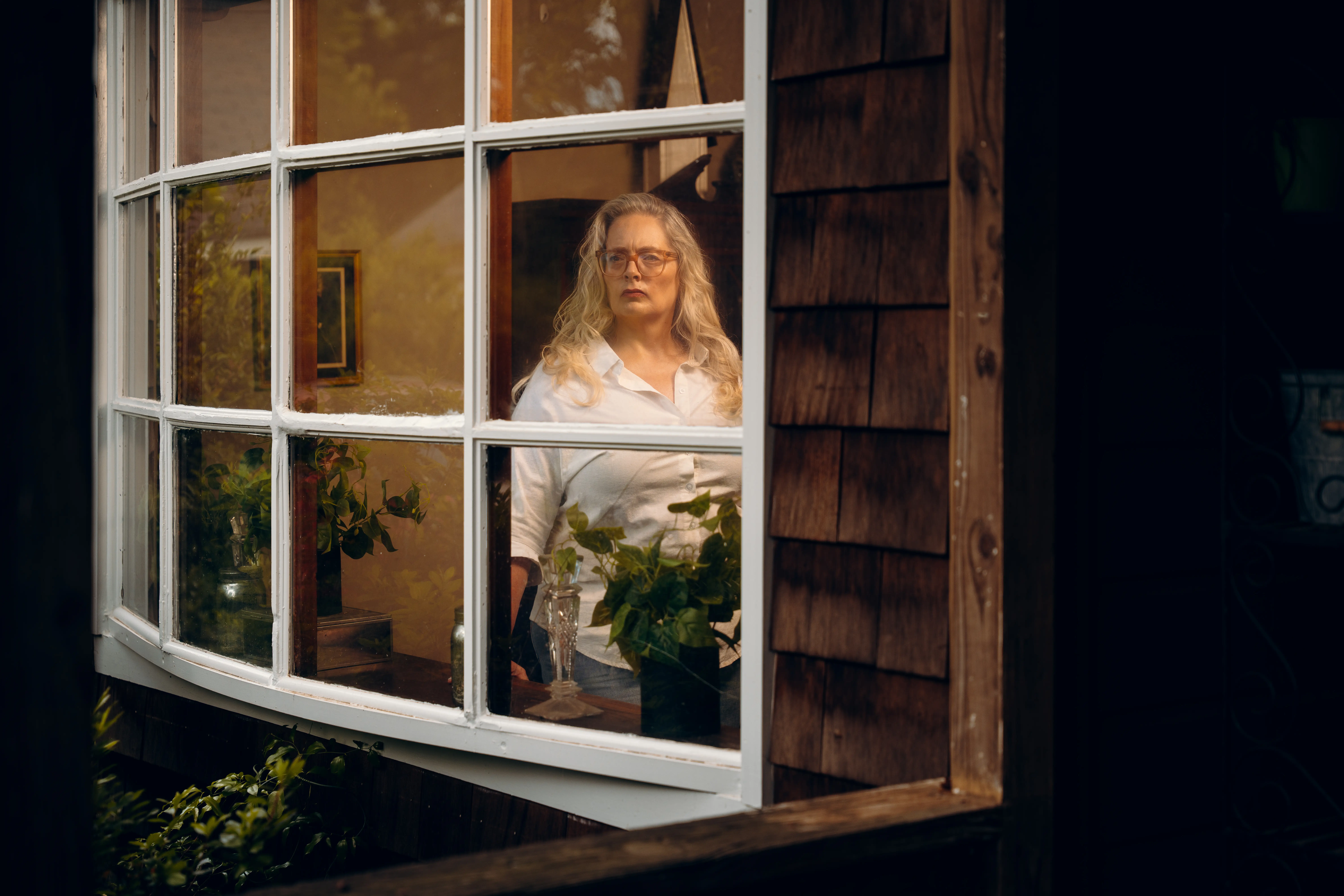 Julie Peck with long blond hair and glasses stands inside a warmly lit room behind a multi-pane window.