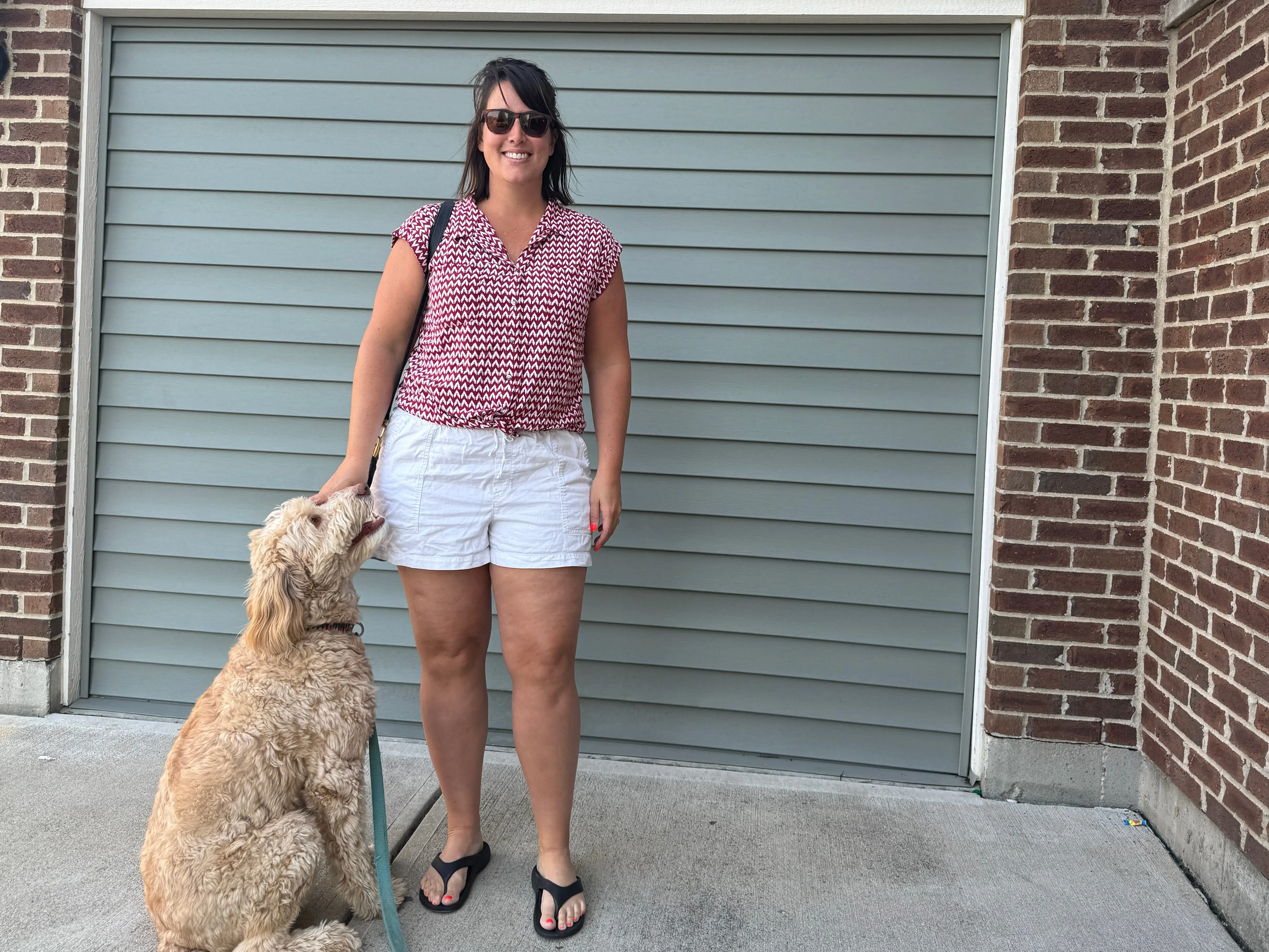 Woman in reddish pink shirt, white shorts,  posing in front of garage and brick wall with dog