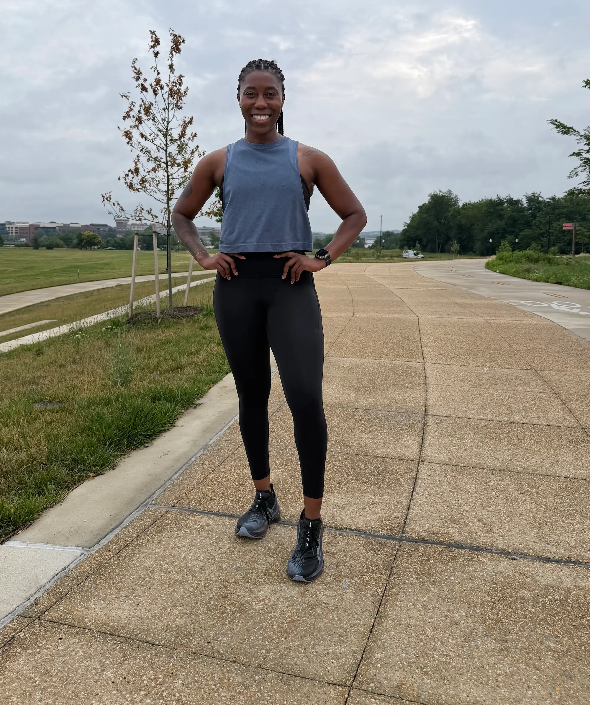Smiling person in athletic wear stands on a paved park path under cloudy skies.