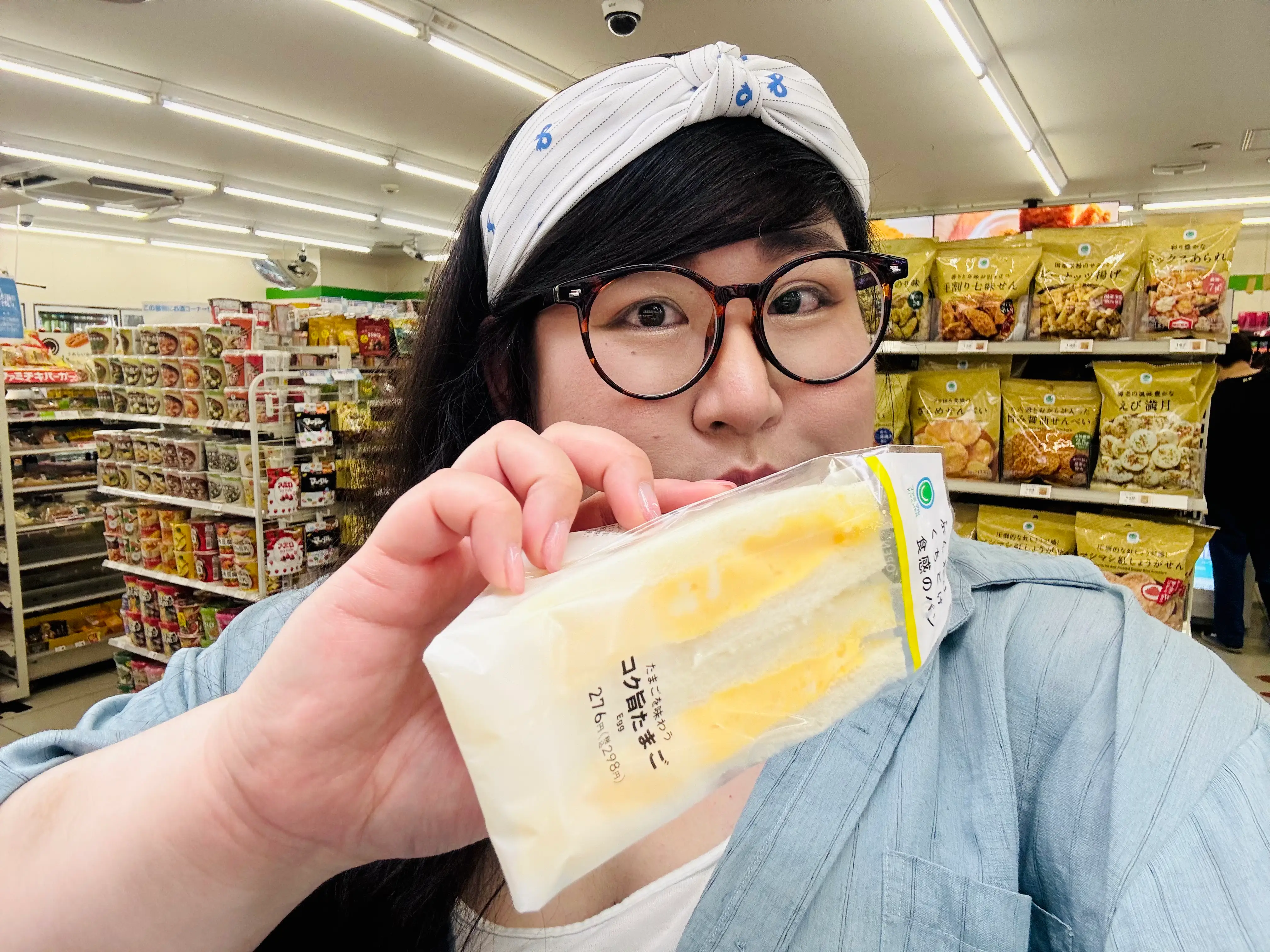 Person wearing glasses holds a packaged sandwich close to the camera inside a brightly lit convenience store.