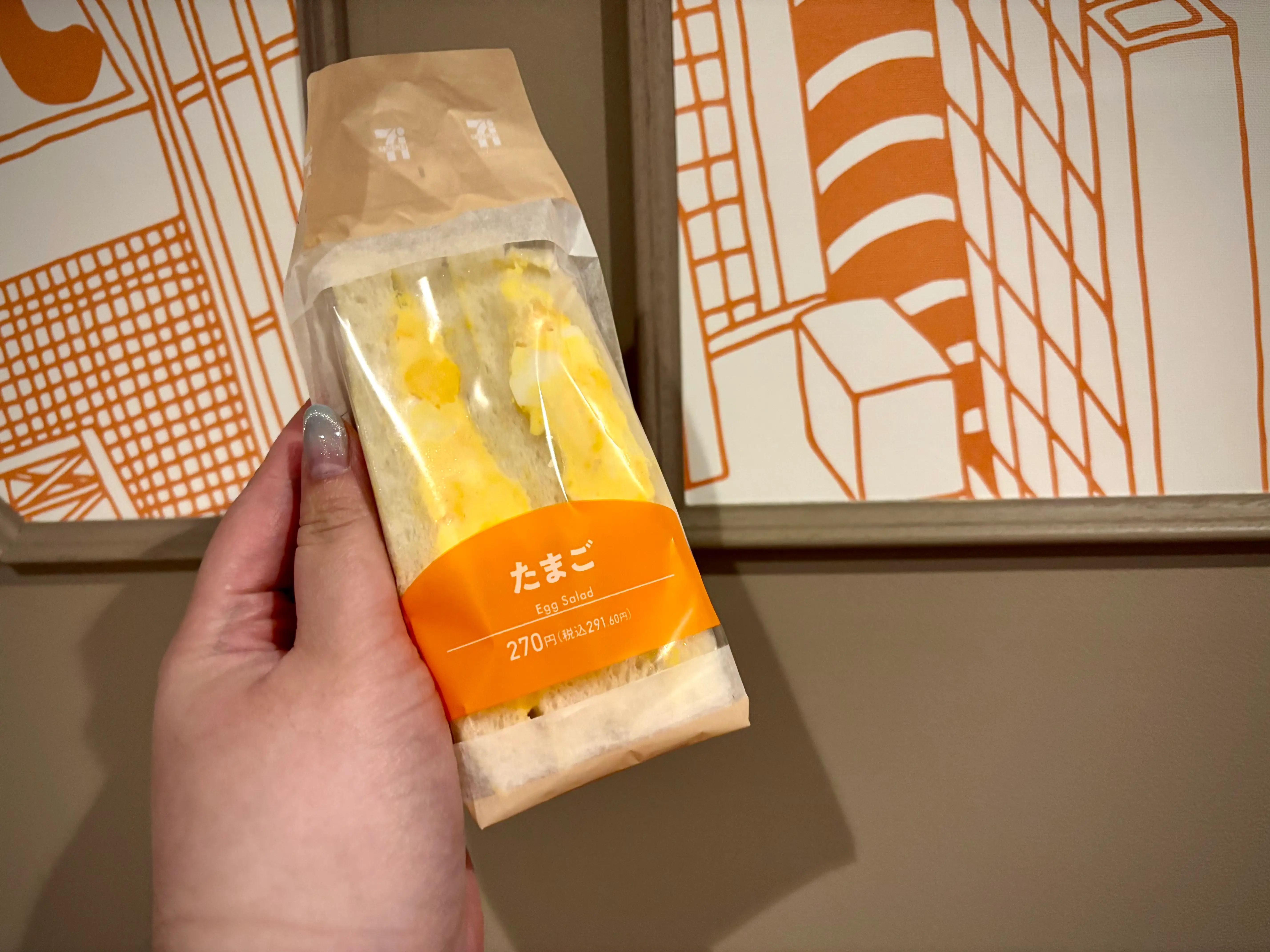 A hand holds a packaged egg salad sandwich labeled たまご against orange line-art wall panels.