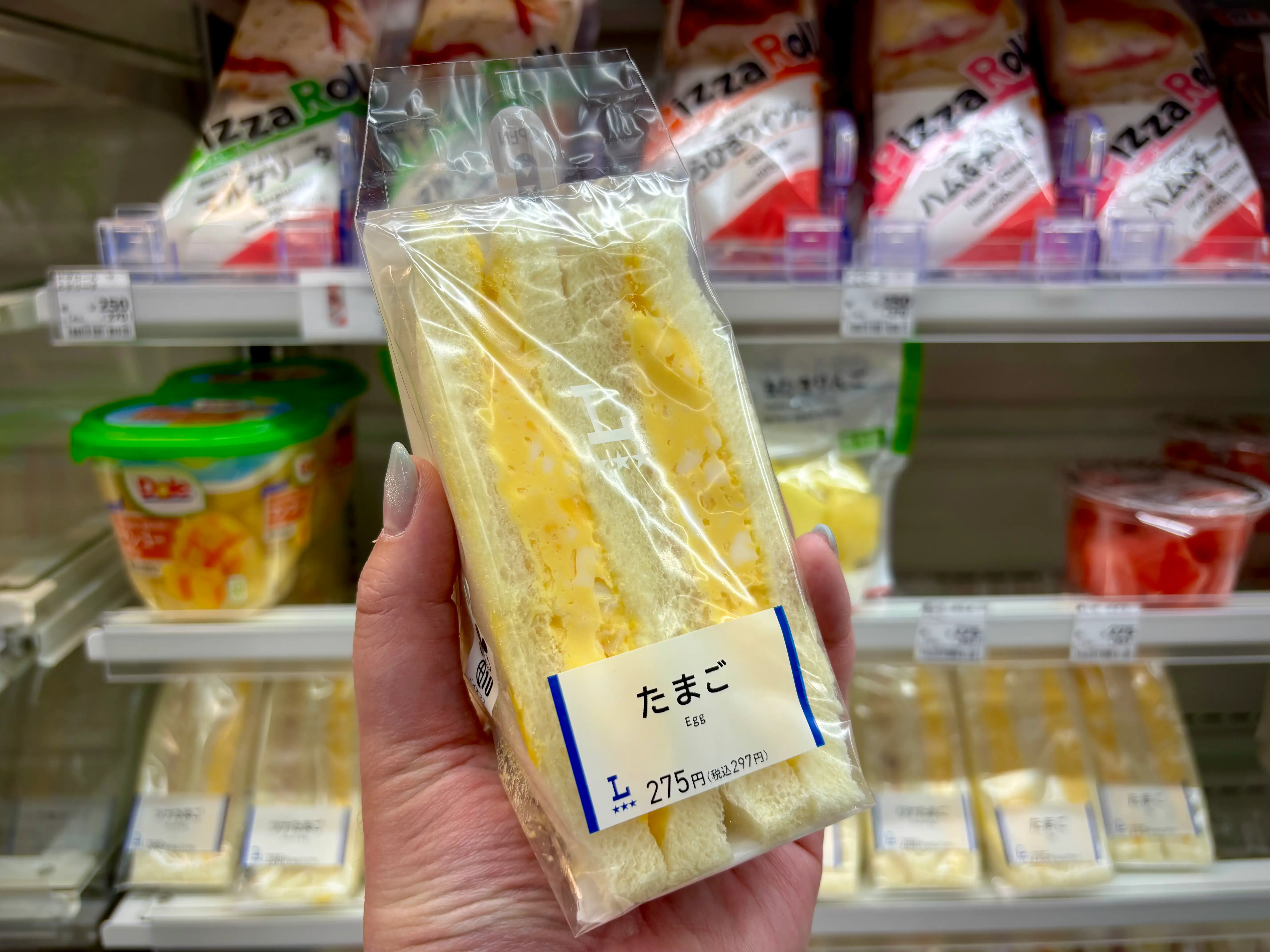 A hand holds a packaged たまご Egg sandwich in front of refrigerated convenience store shelves.