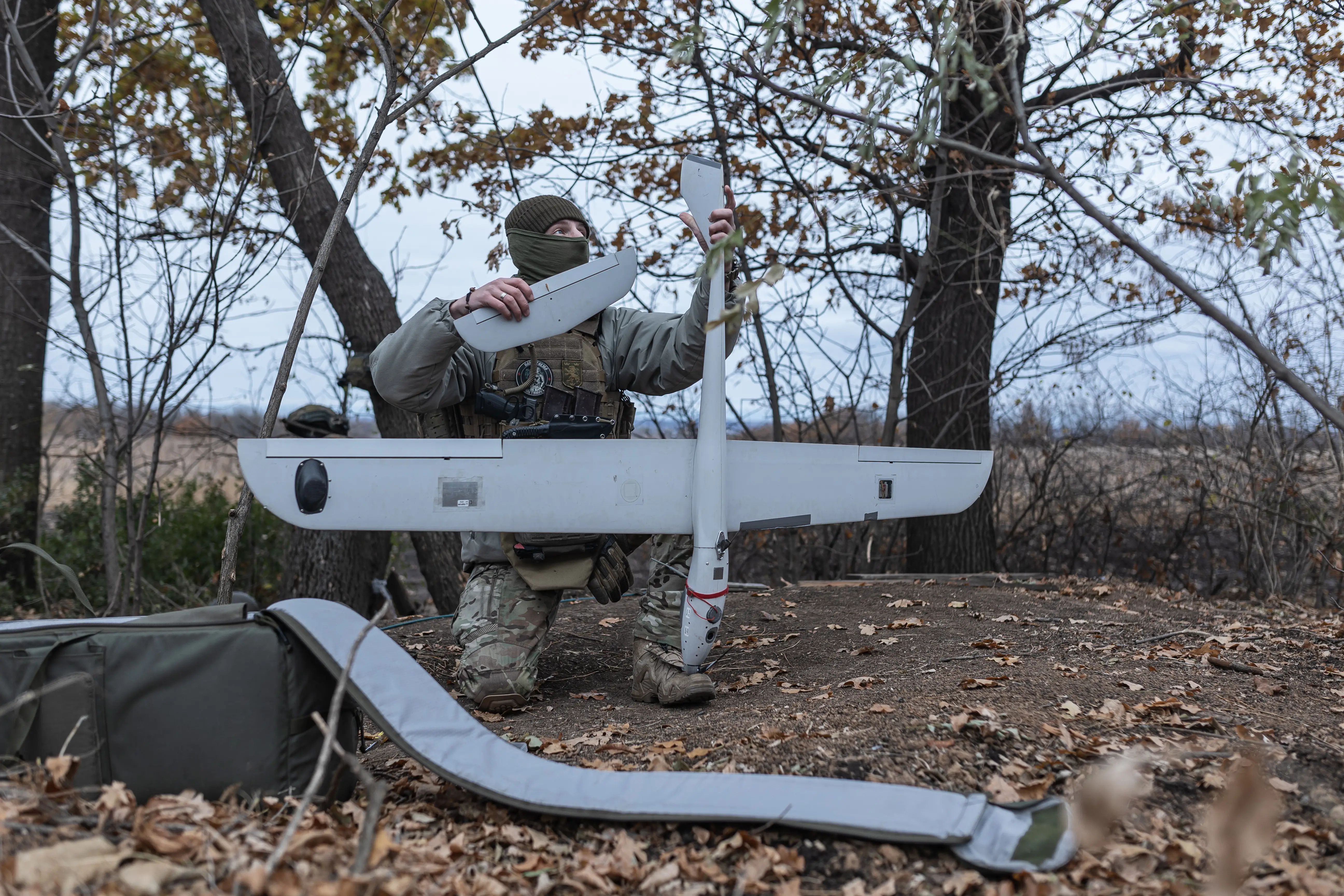 A man in camoflage gear and a face covering knees in a muddy field with bare trees working on a large grey drone