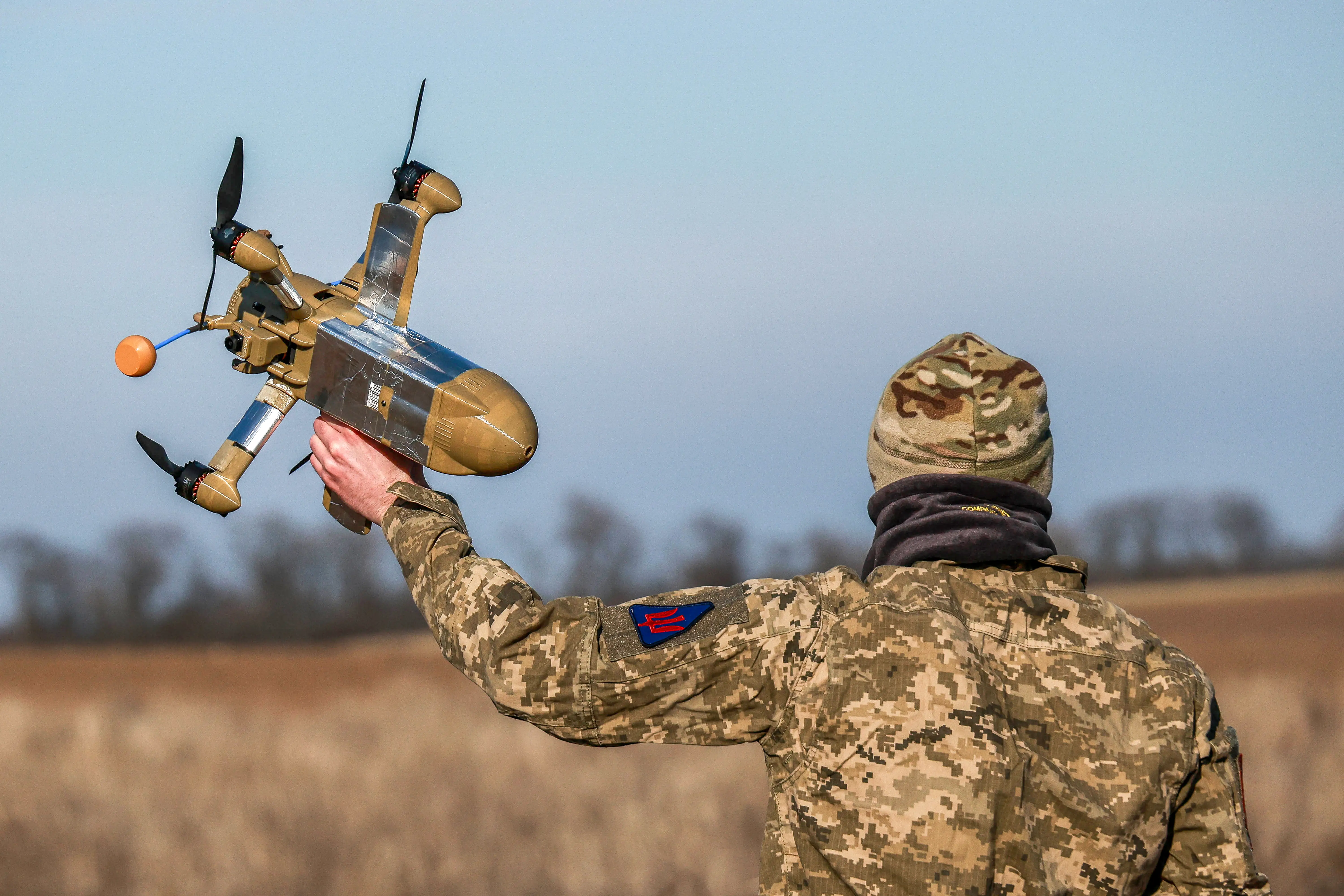 A man in a camouflage top and beanie stands with his back turned in a field holding a khaki and silver drone in the air with one hand