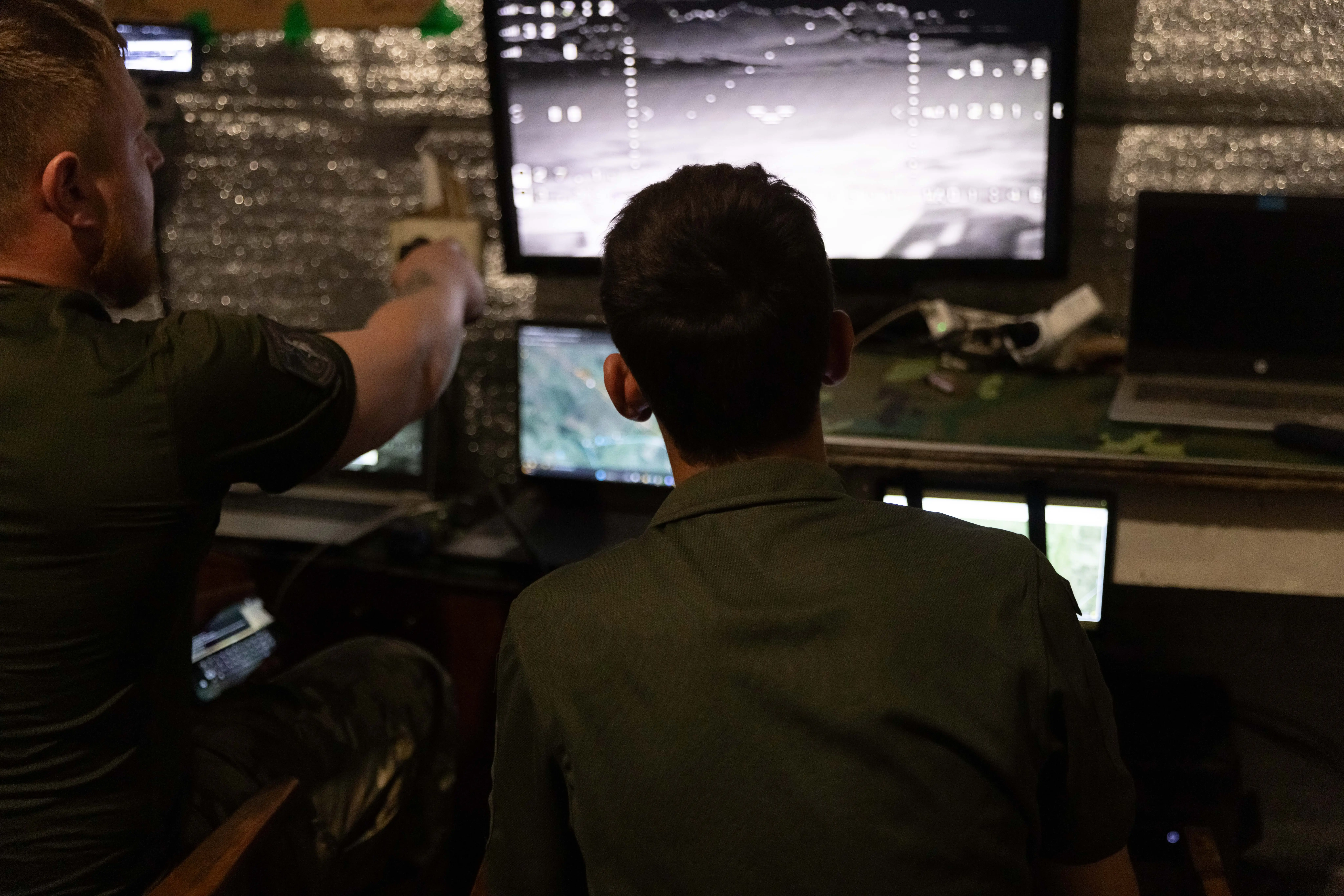 Two men sit with their backs turned looking at a screen that shows a black-and-white drone feed