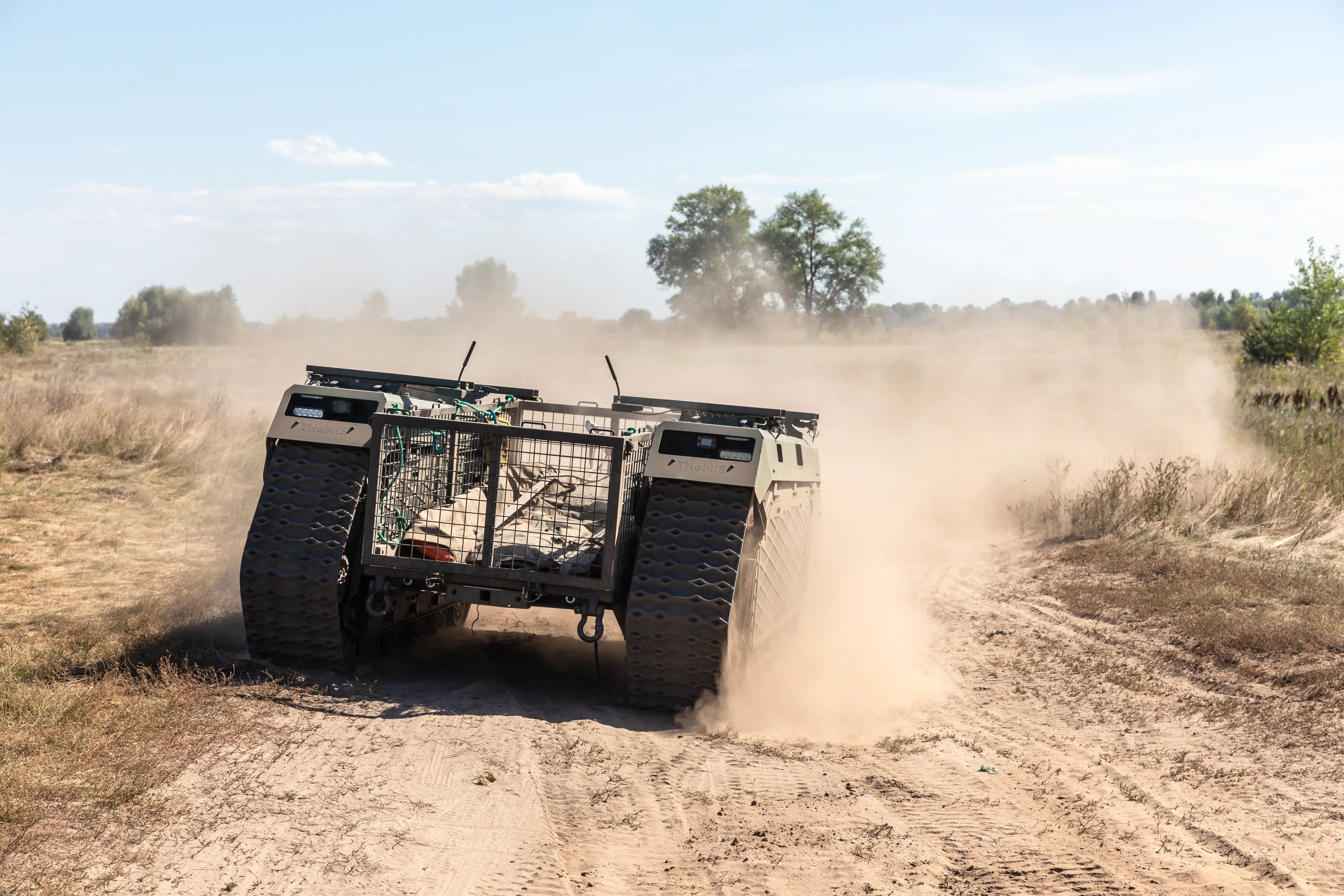 A robot on tracks drives along a pale, dusty track with dust in the air a trees and blue sky behind them