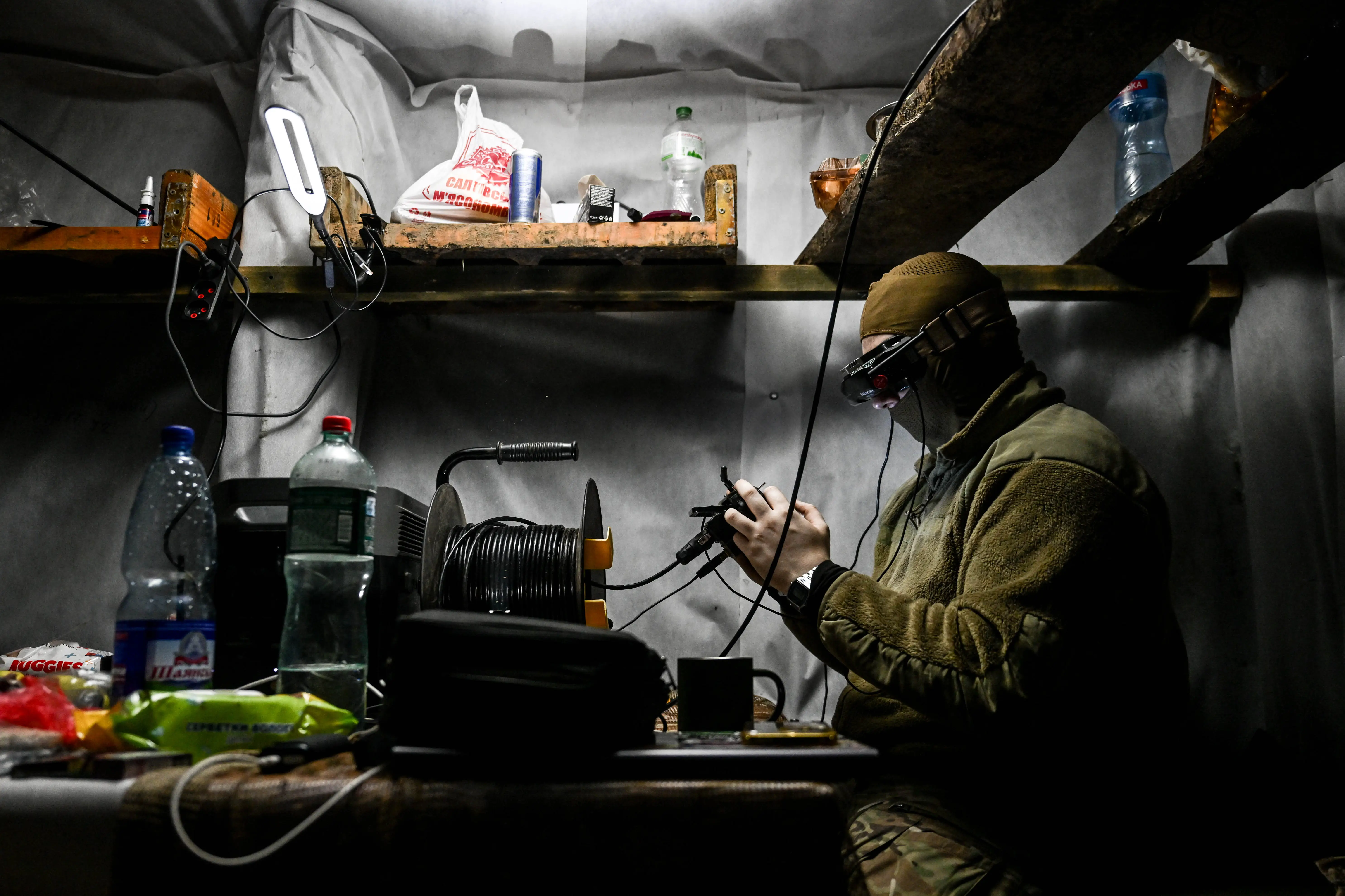 A man wearing khaki and a hat sits in a small indoor location with a headset and a controller surrounded by water bottles and other supplies