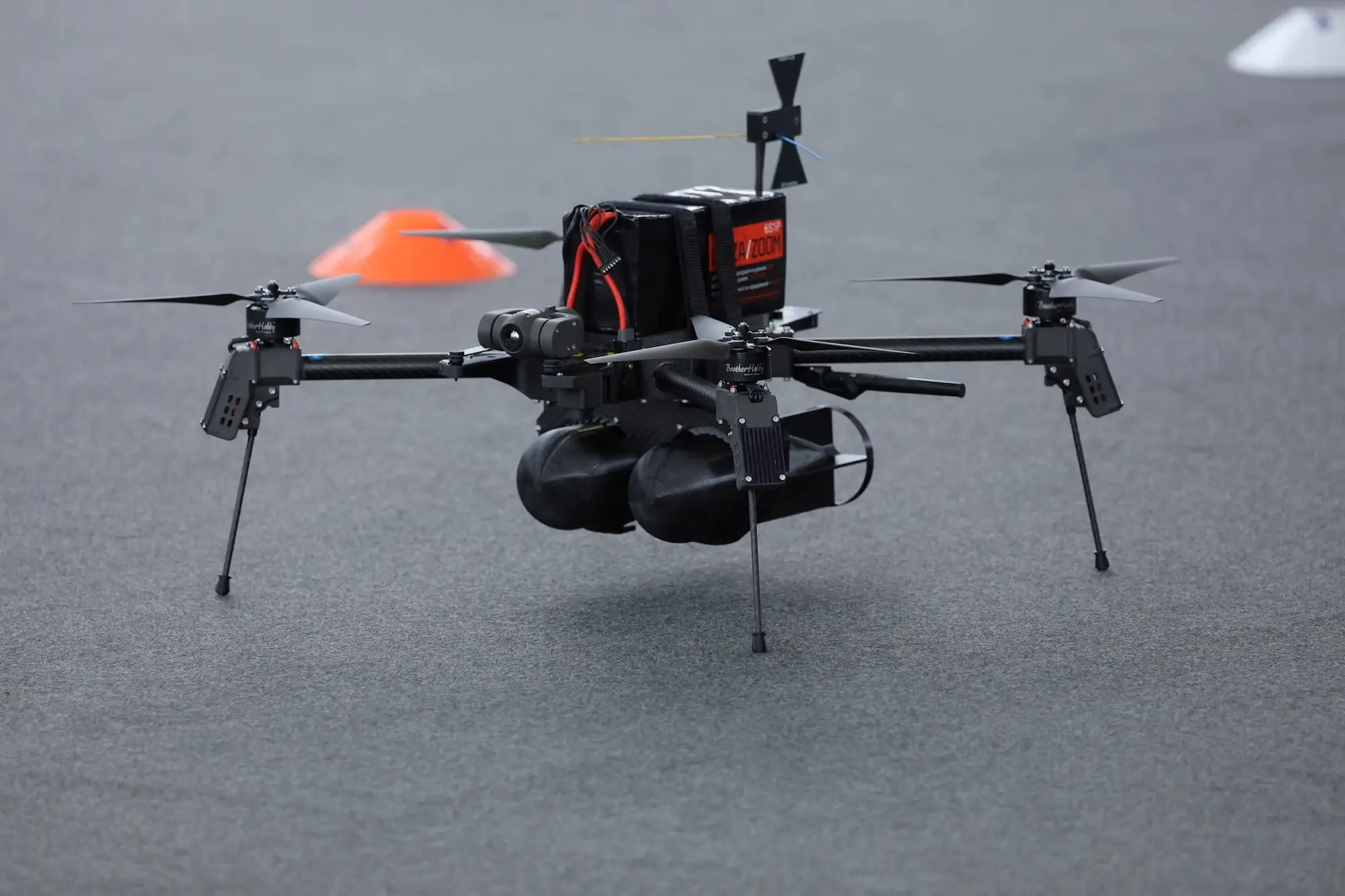 A black quadcopter drone resting on grey tarmac with a small orange cone on the ground