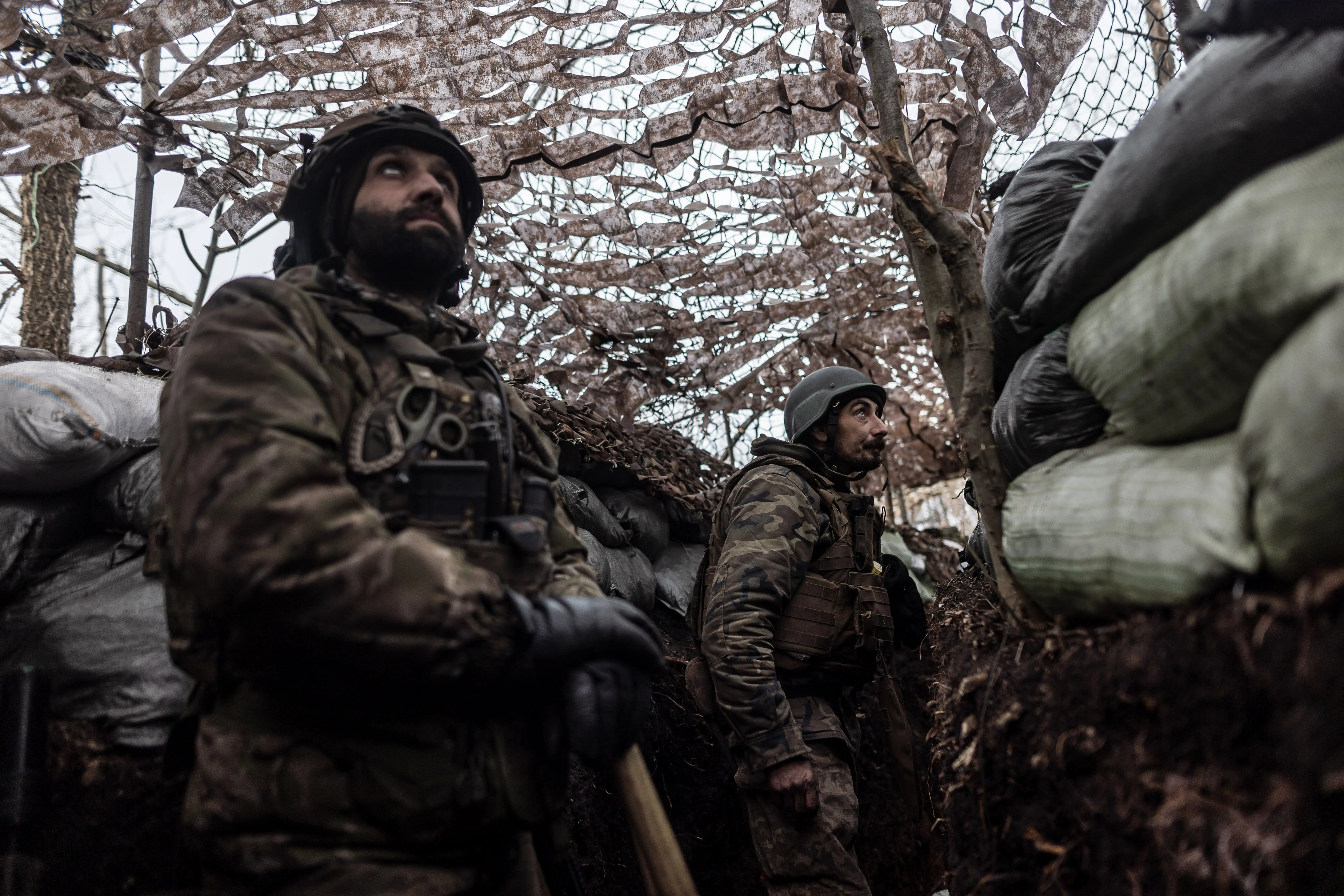 Two men in camouflage hear and helmets stand in a sandbag-lined position under a net, looking up