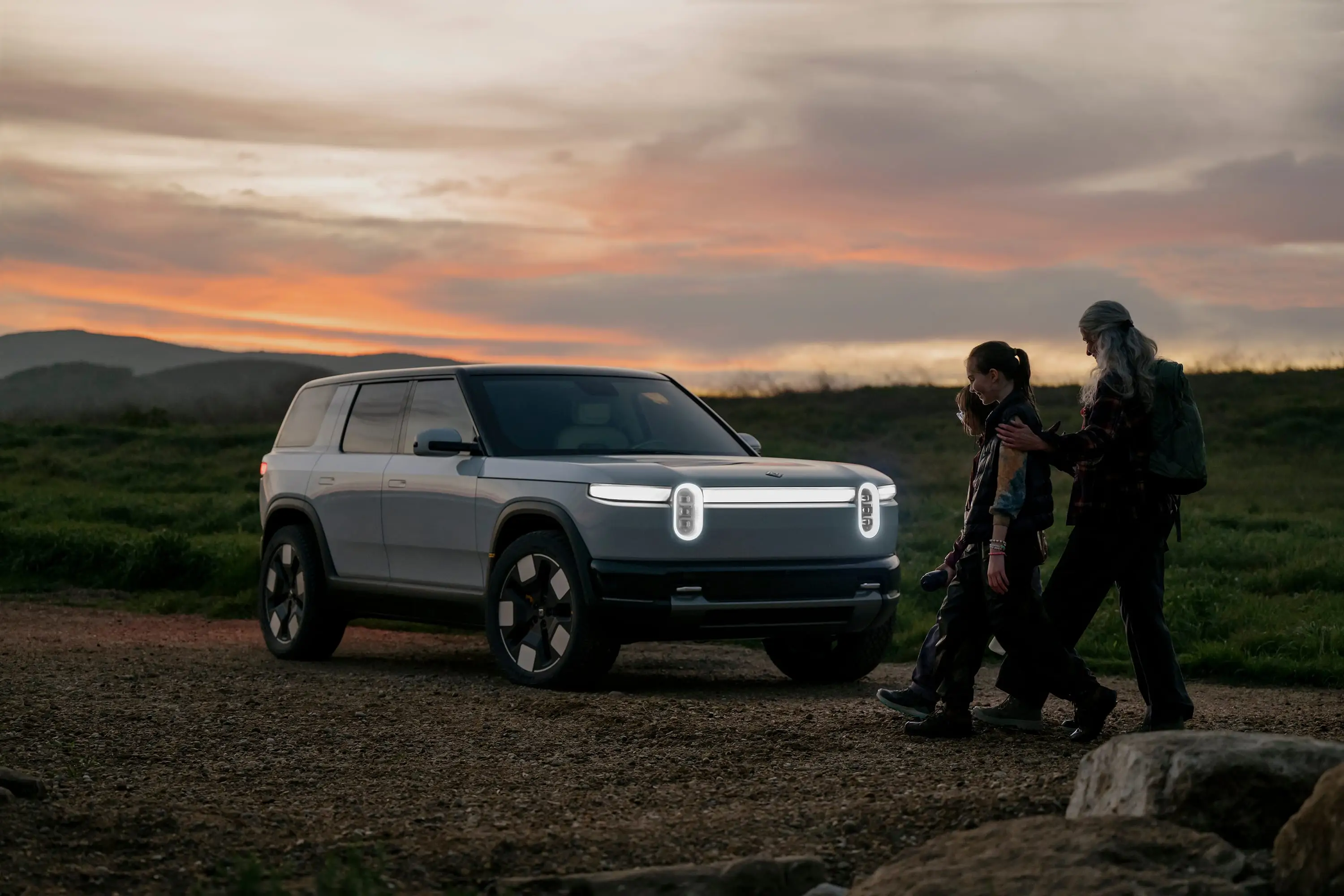 Three people walk toward a white Rivian R2 that's parked on gravel.
