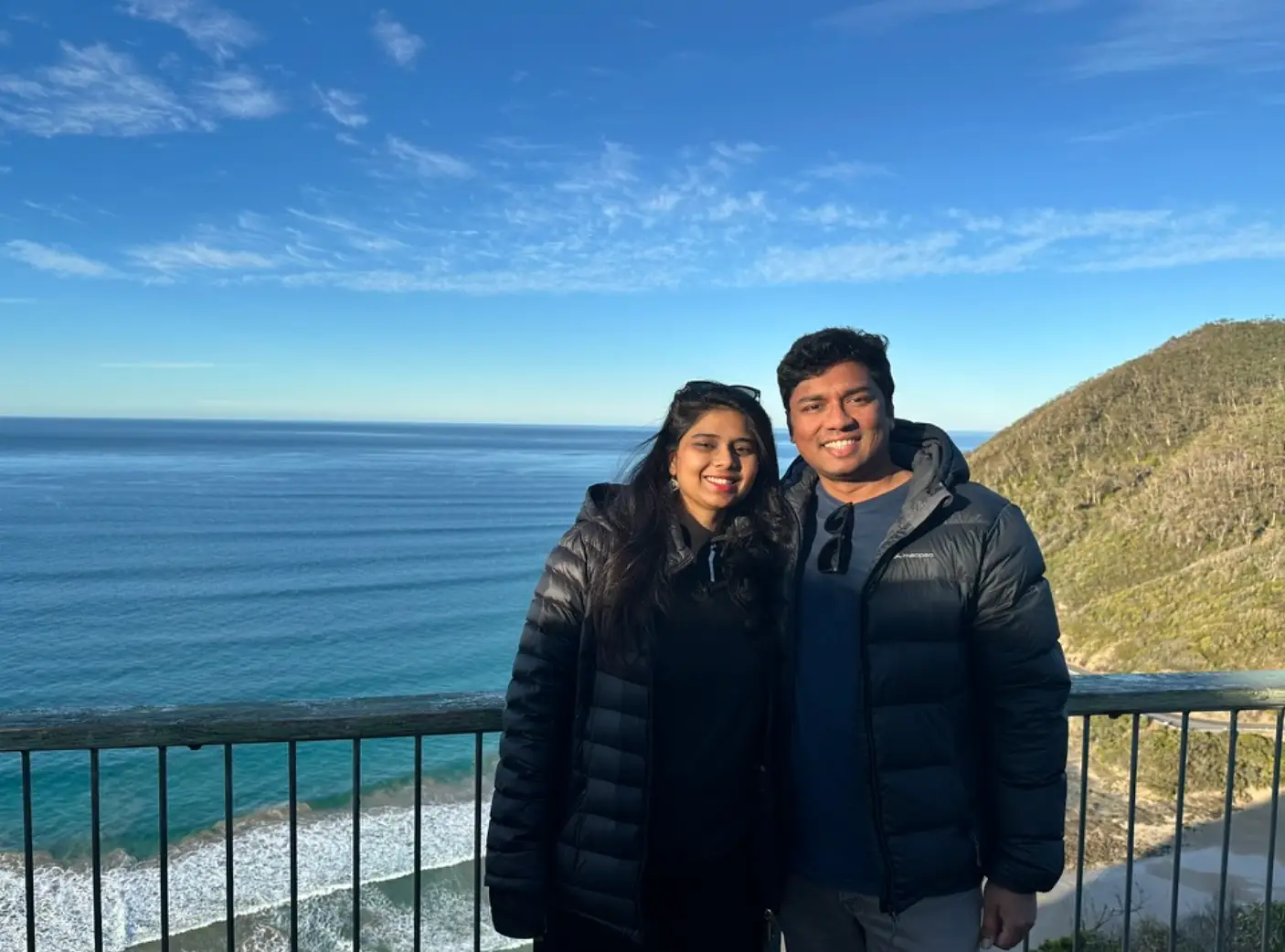 The writer and her husband wearing black puffer coats and standing in front of a beach view on a trip.
