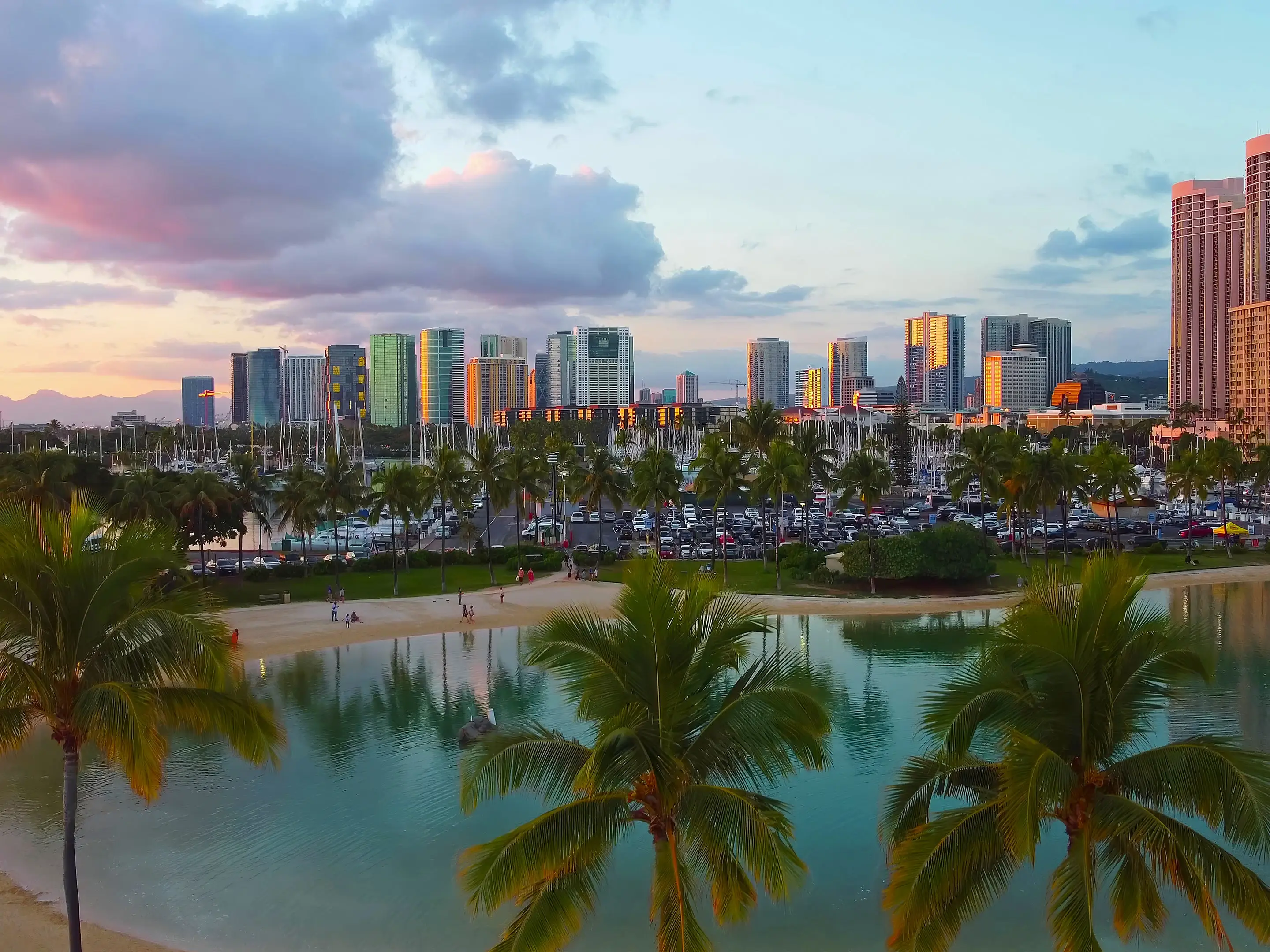 Honolulu skyline and lagoon at sunset. Scenic view of a tropical lagoon with palm trees, sandy beach, and the illuminated Honolulu skyline reflecting the warm hues of sunset