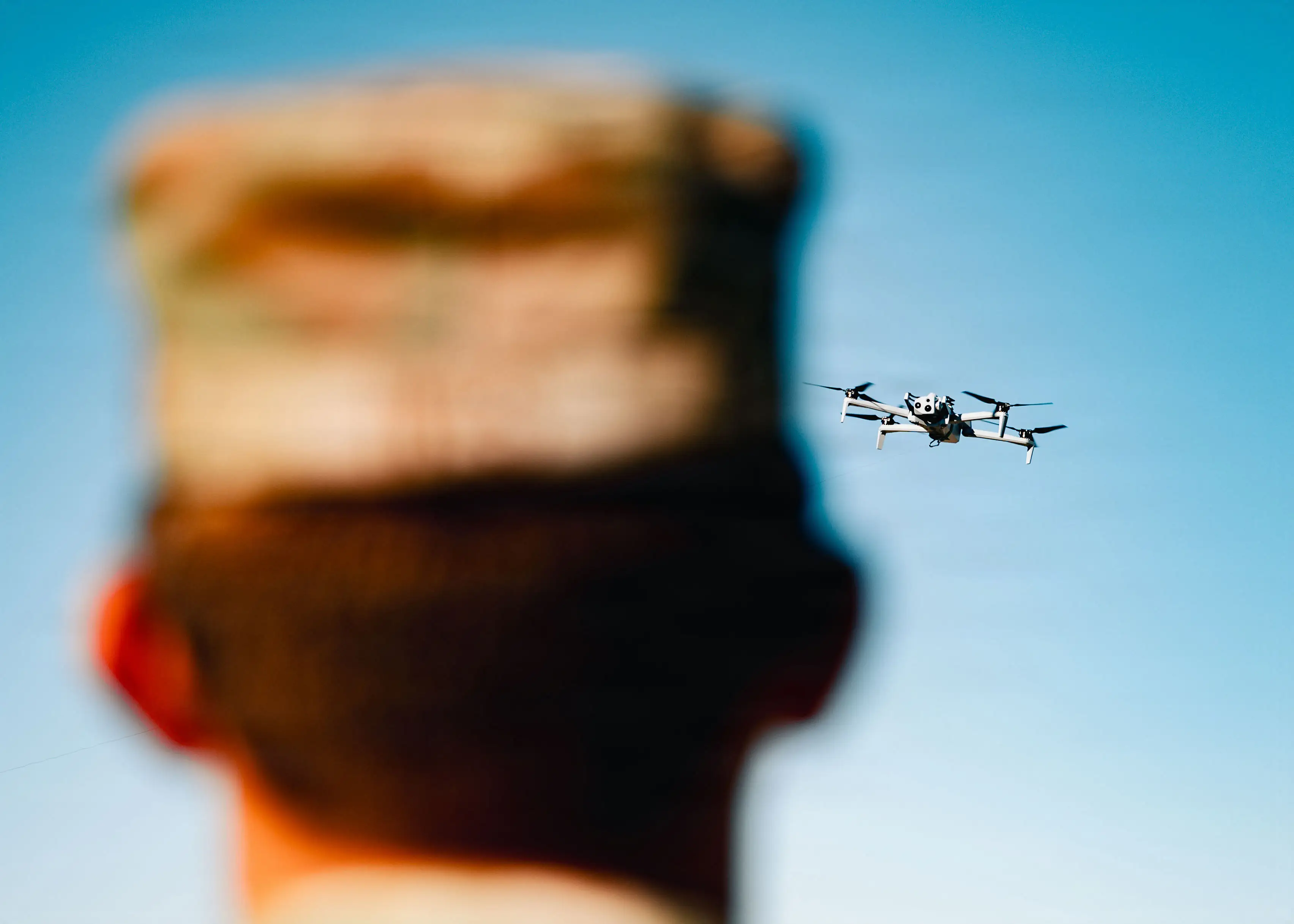 A small quadcopter drone flies against a clear blue sky. The back of a man's head is seen in the foreground.