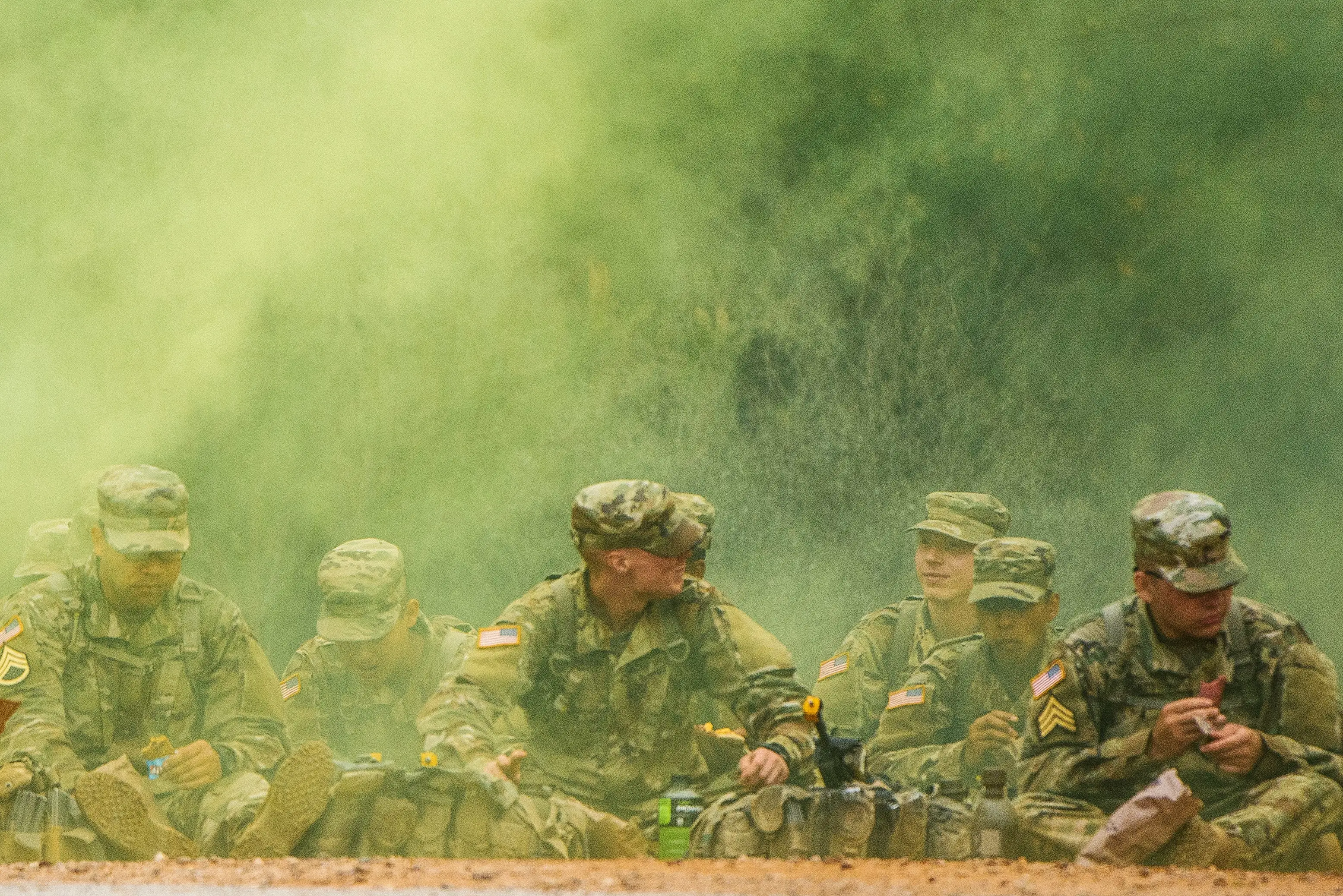 Camouflage-clad military personnel sit behind a barrier amid a dense cloud of green smoke during an outdoor exercise.