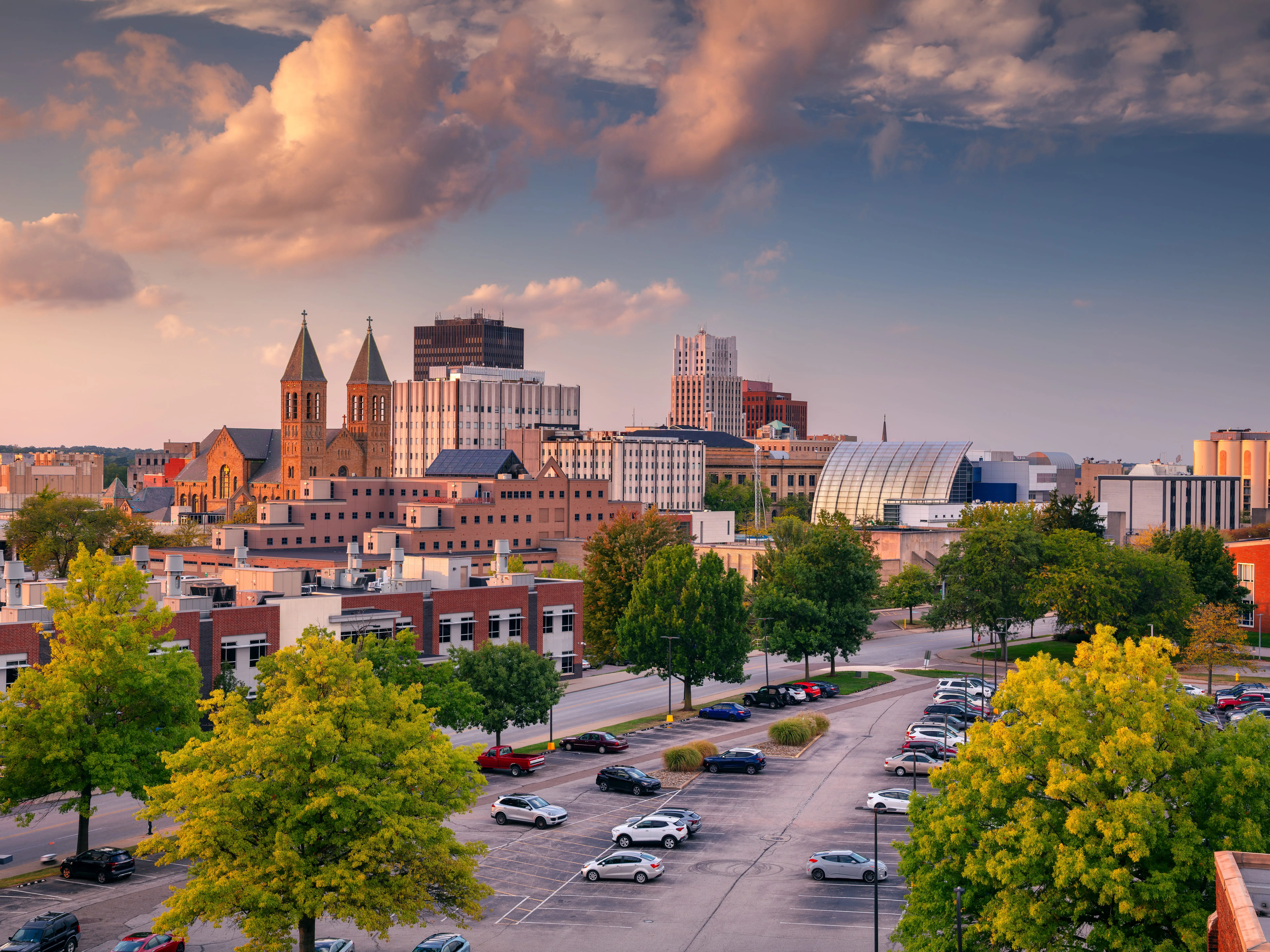 Downtown Akron, Ohio, USA at autumn sunset features a mix of modern and historic architecture. Prominent are twin-spired churches and contemporary buildings. The foreground shows a parking lot with evenly spaced cars and vibrant green trees. In the background, the skyline rises under a dramatic clouded sky, colored in warm hues by the setting sun. The scene captures a serene and picturesque moment in the city.