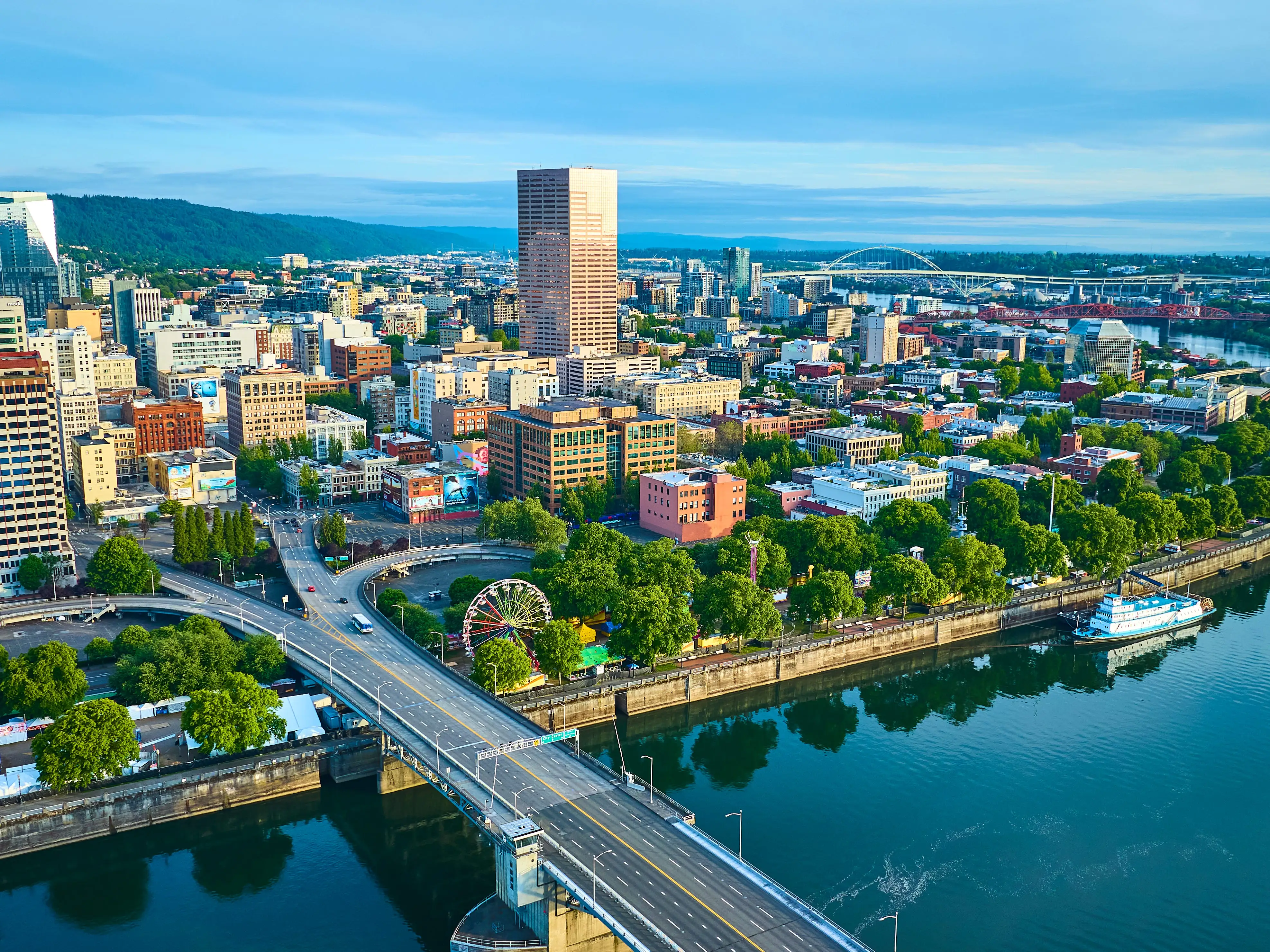 Aerial view of Portland, Oregon, showcasing the vibrant downtown with a mix of modern and classic skyscrapers. The Willamette River with a bridge and paddlewheel riverboat adds scenic beauty and historical charm.