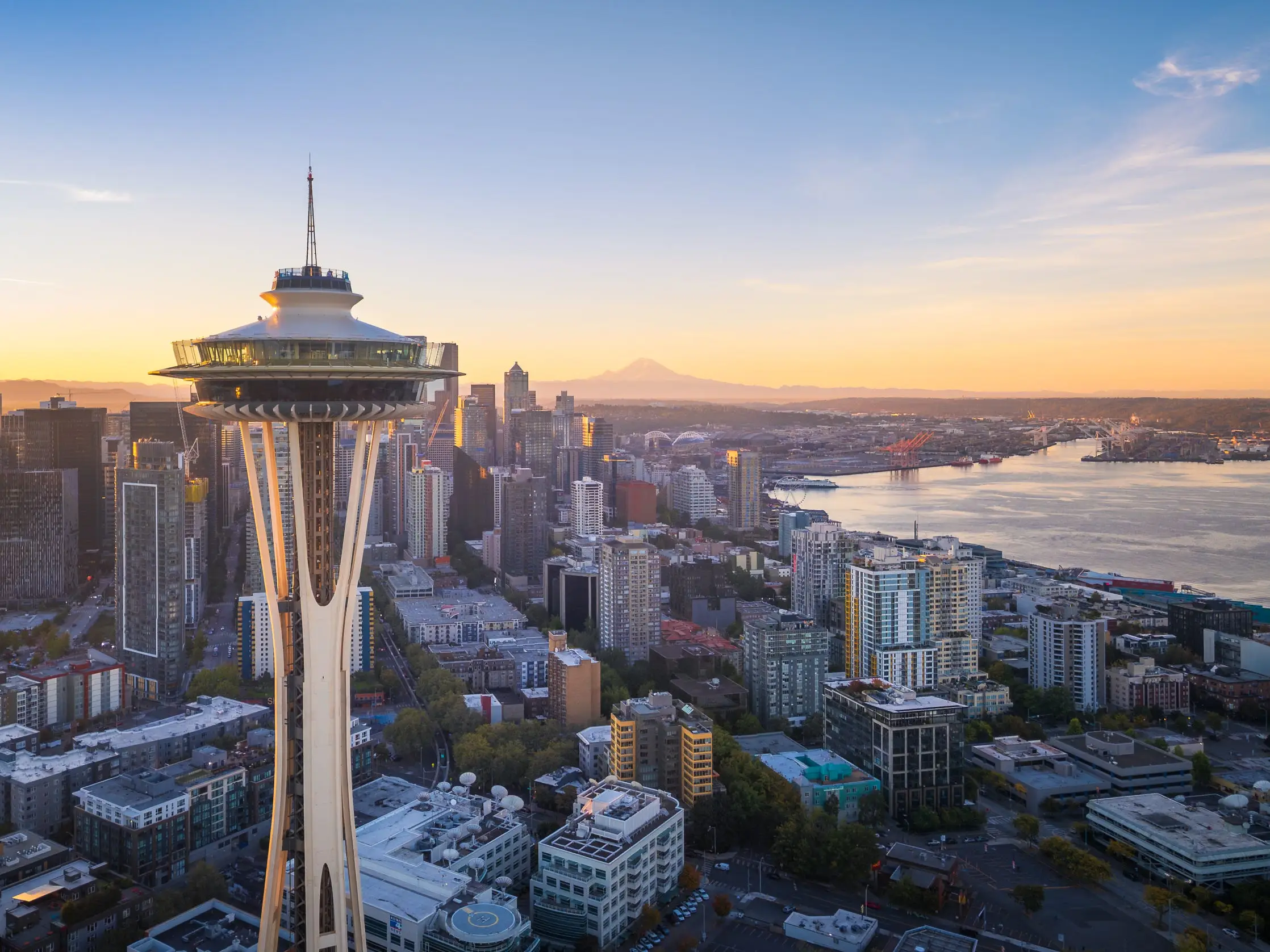 Aerial view of Seattle skyline with the Space Needle in the foreground and Mount Rainier rising in the distance. Clear skies highlight the city's modern architecture and natural beauty.