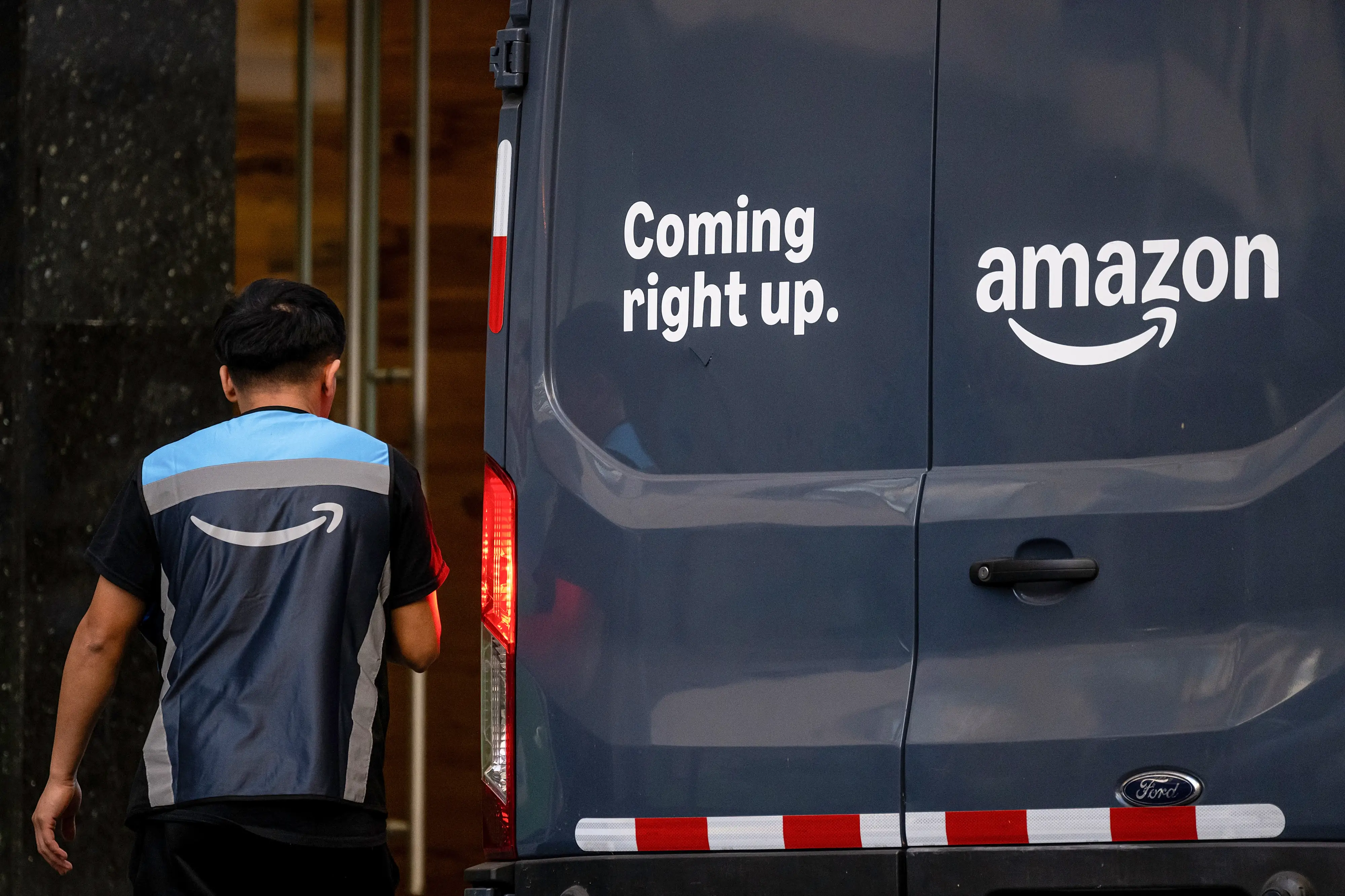 An Amazon delivery worker wears a blue-and-black vest while walking past the back of a black Amazon delivery van with the Amazon name and 
