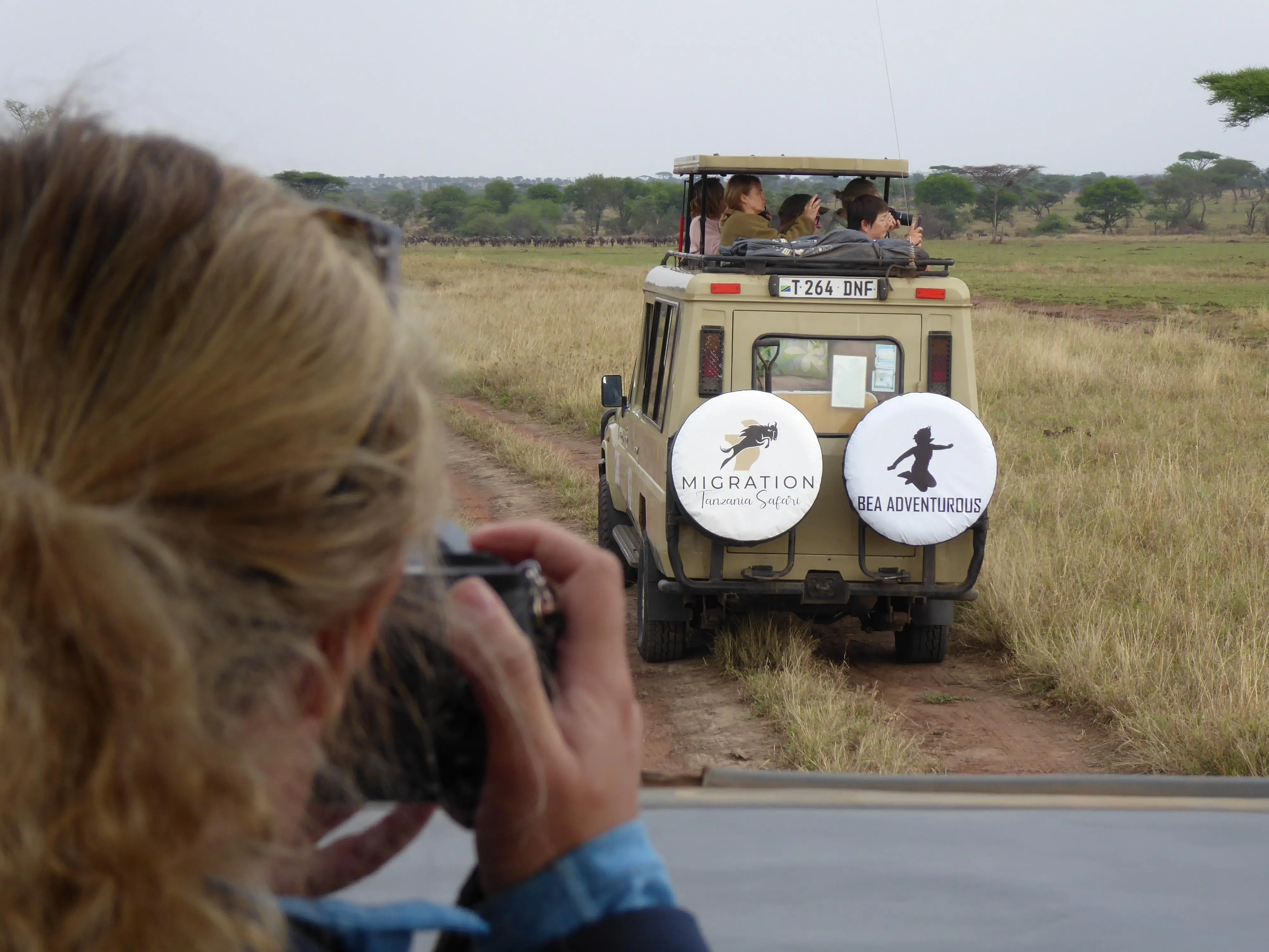 A woman on safari takes a picture with a safari vehicle in front of her.