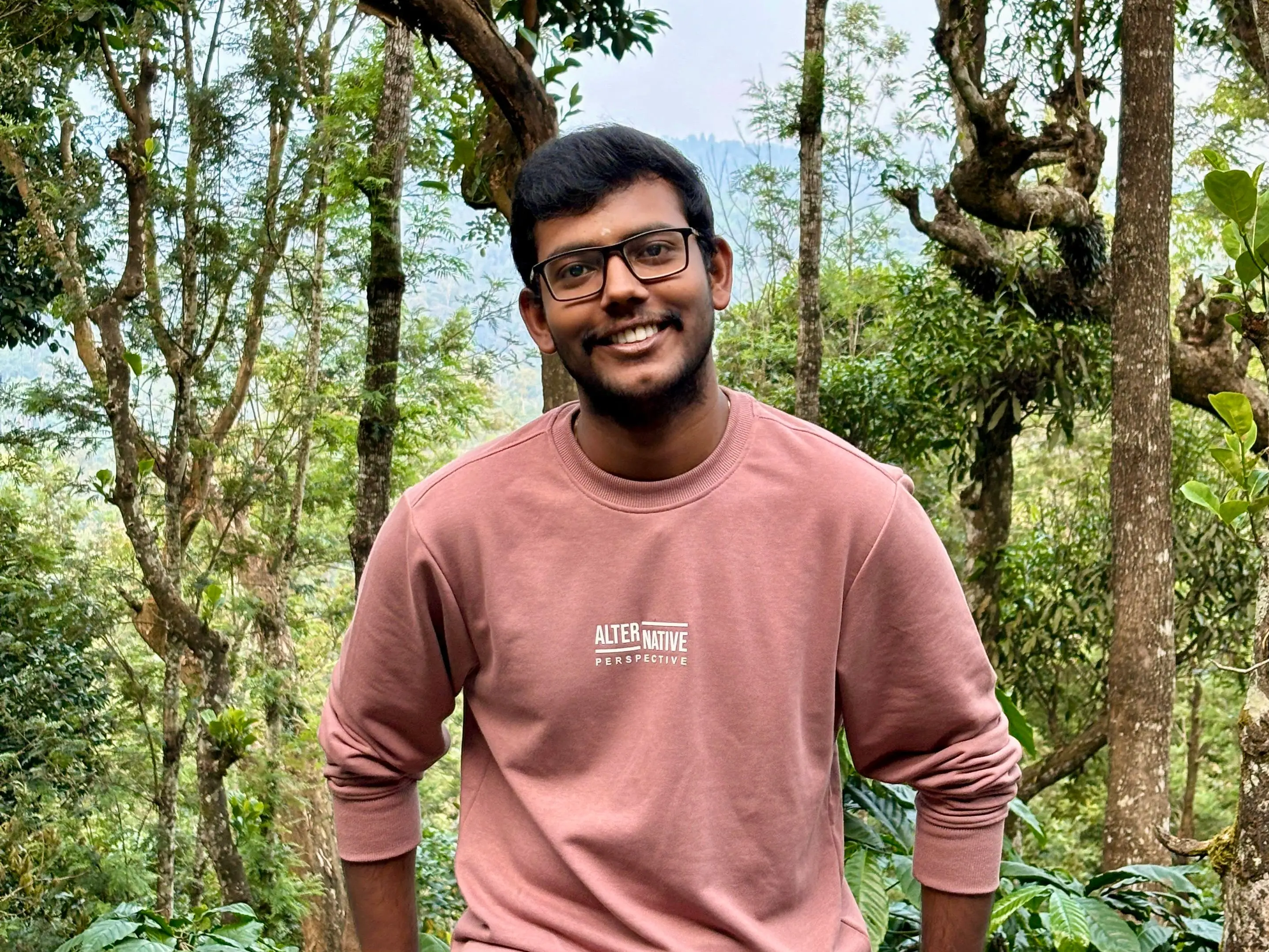Rathesh Prabakar is wearing a pink sweatshirt and standing in front of some trees.