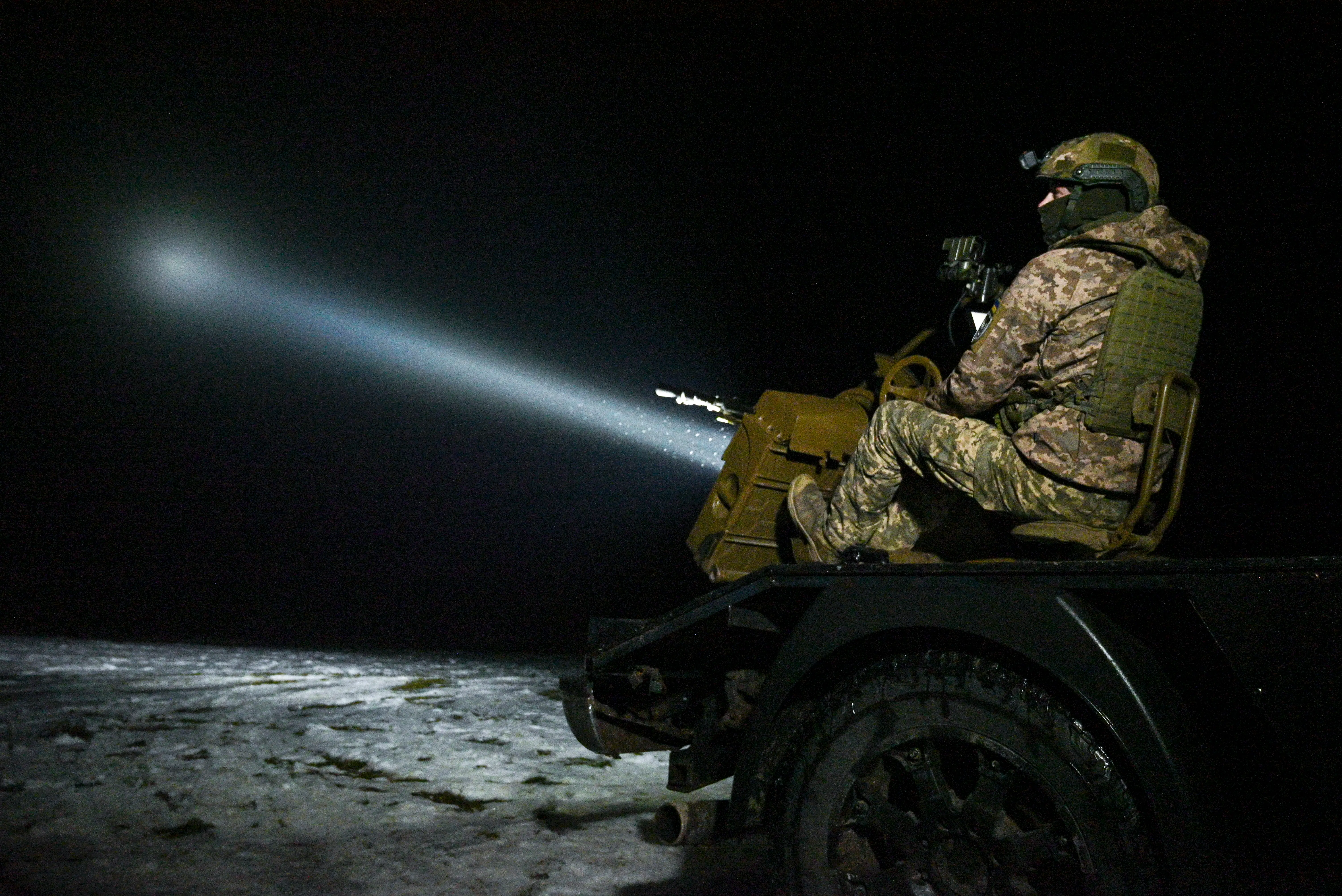 A man in camouflage gear and a helmet sits on the back of a black vehicle under a dark sky with a beam of white light going into the sky