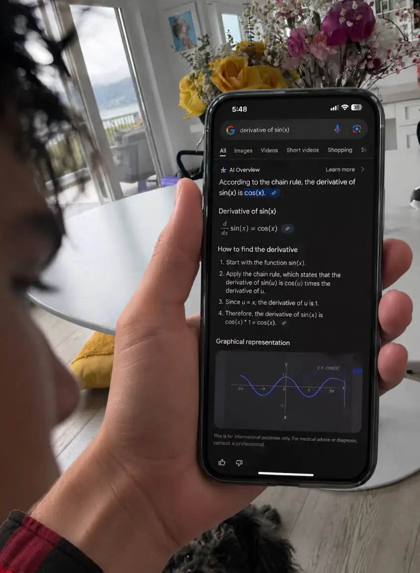 Hand holding a smartphone showing a Google AI Overview for the derivative of sin(x) in a bright room.