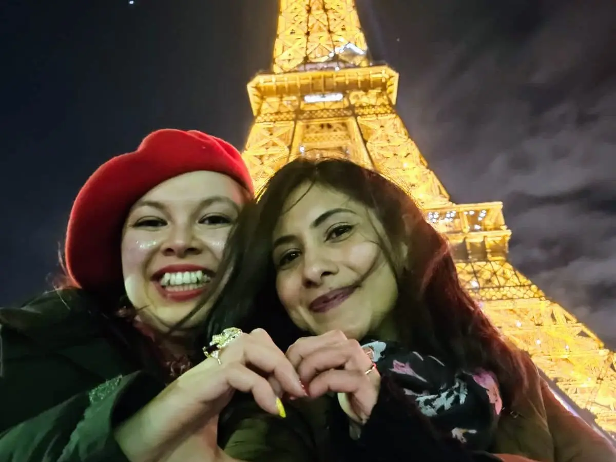 The writer and a friend posing with heart hands in front of the Eiffel Tower at night.