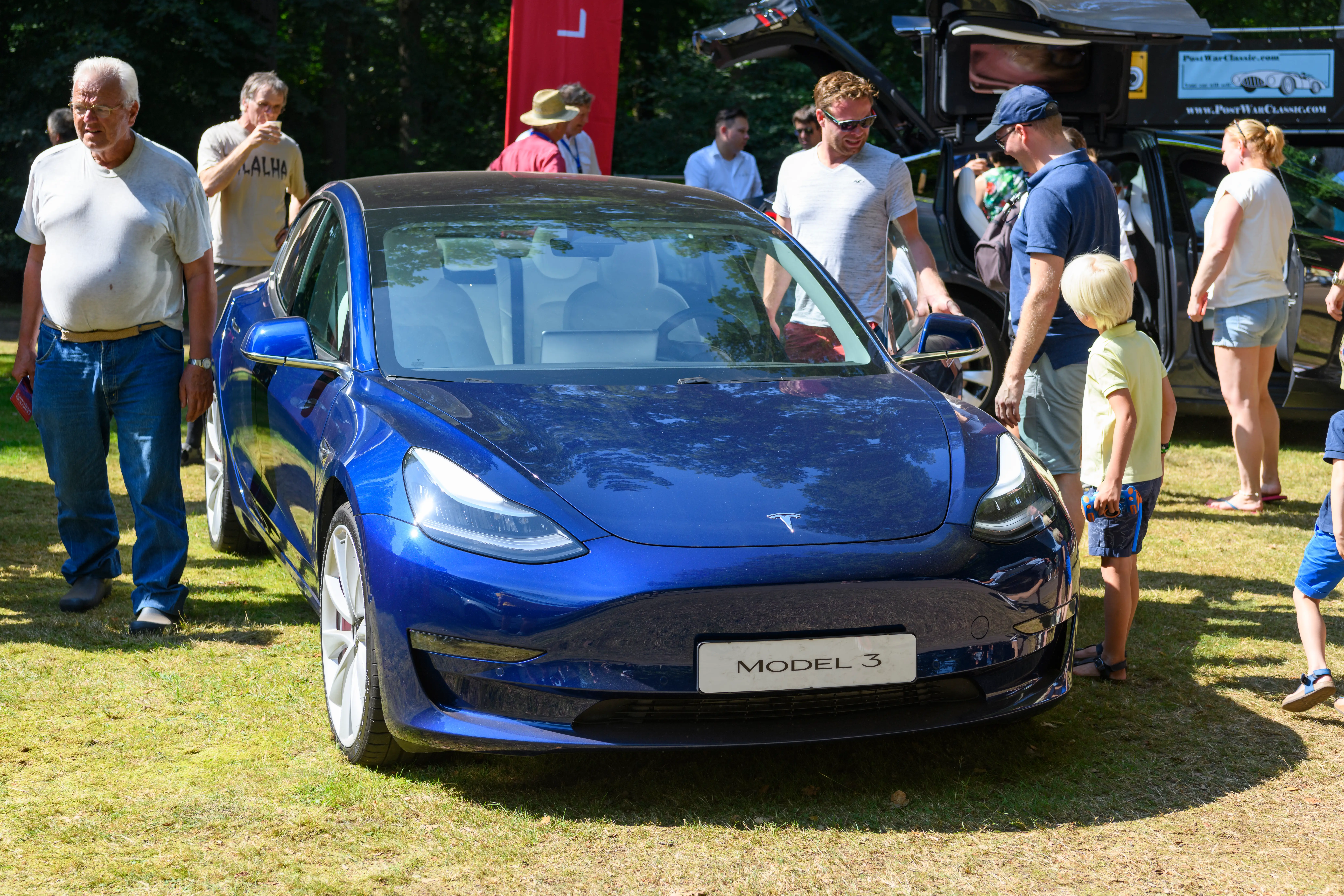 Several people walk around a blue Tesla Model 3 sedan.