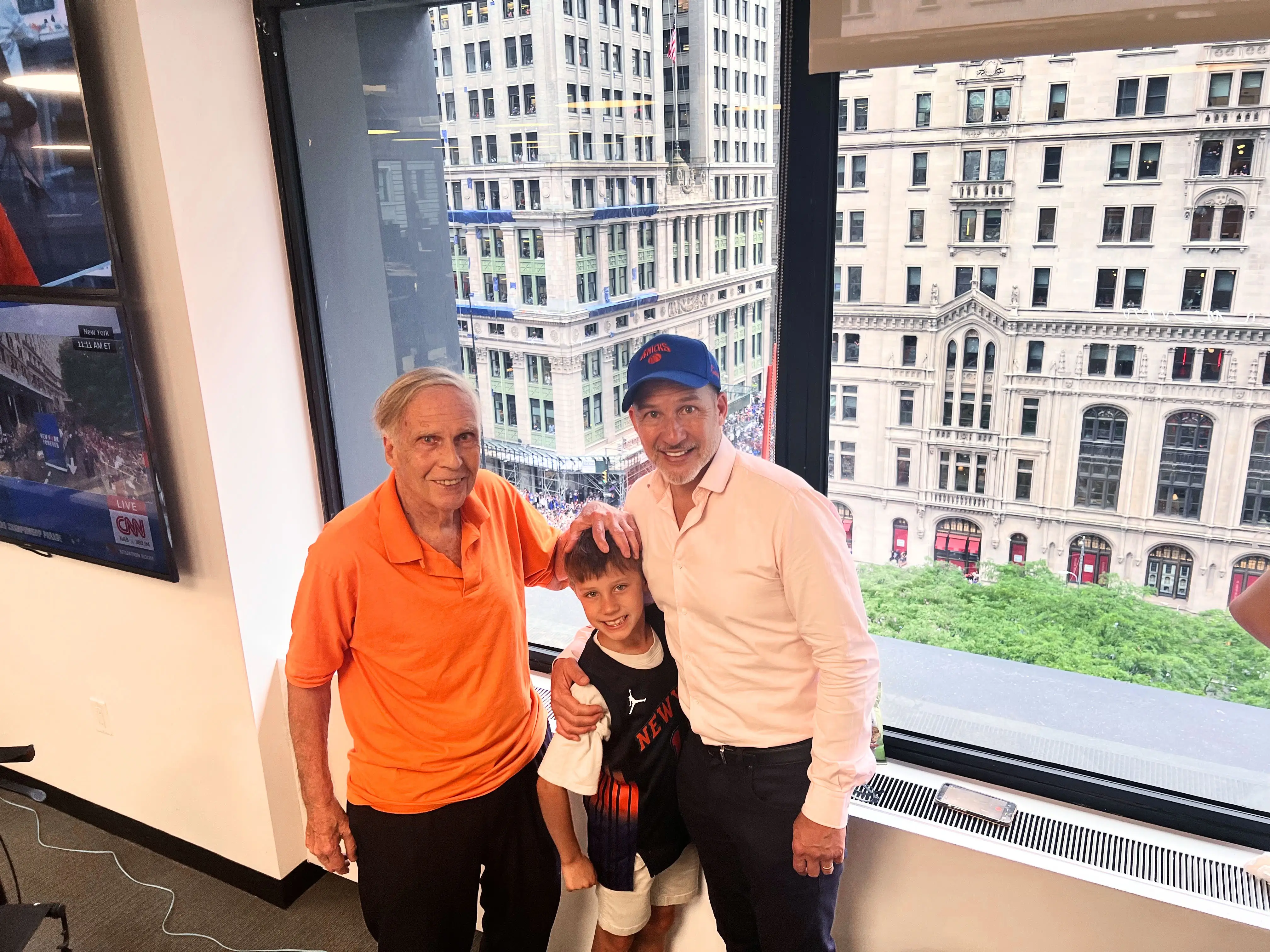An older man, a boy in a New York Knicks jersey, and a man in a blue cap pose by office windows overlooking city buildings.