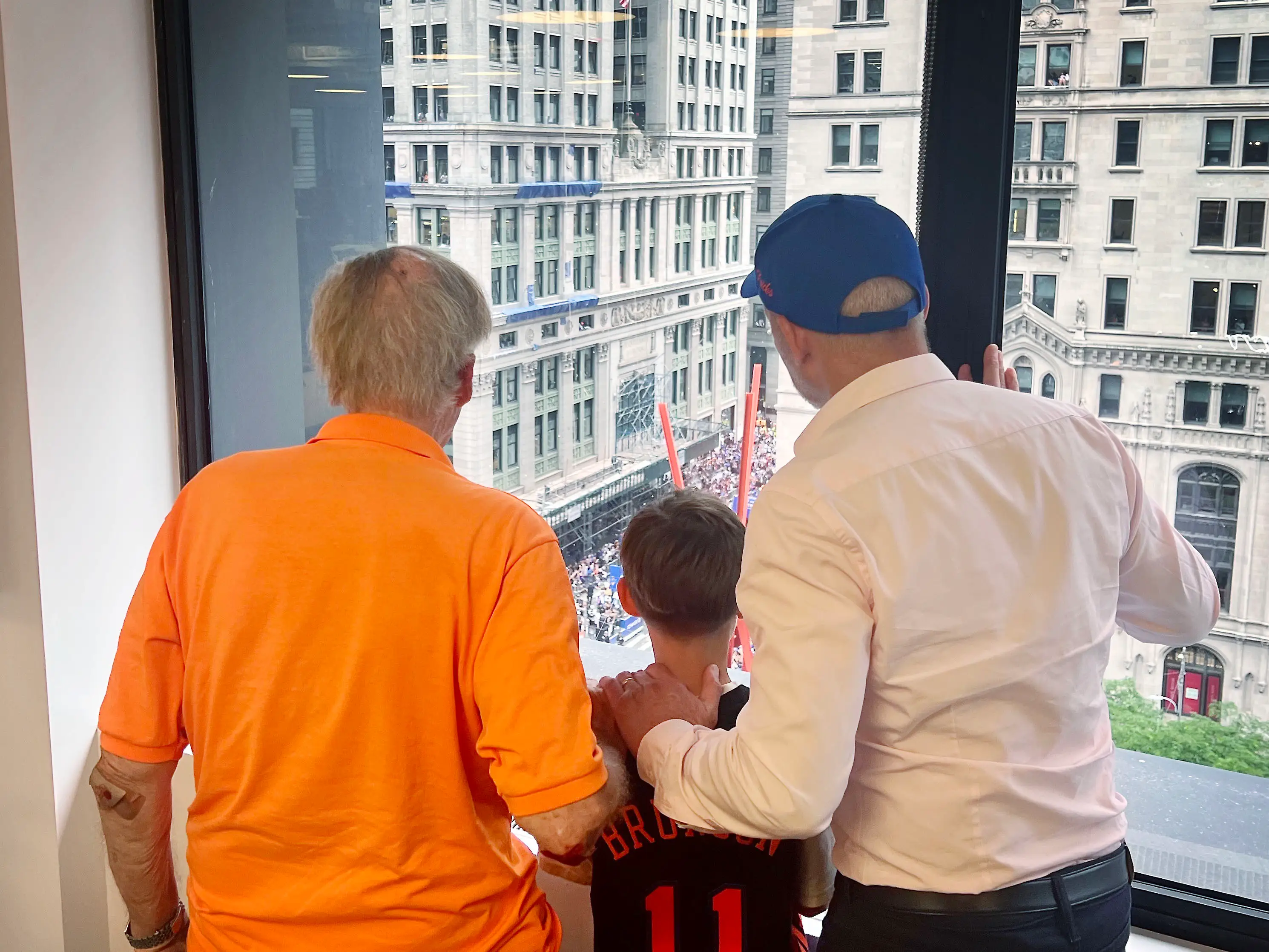 An older man, a boy in a New York Knicks jersey, and a man in a blue cap look out a high window at a crowded street below.