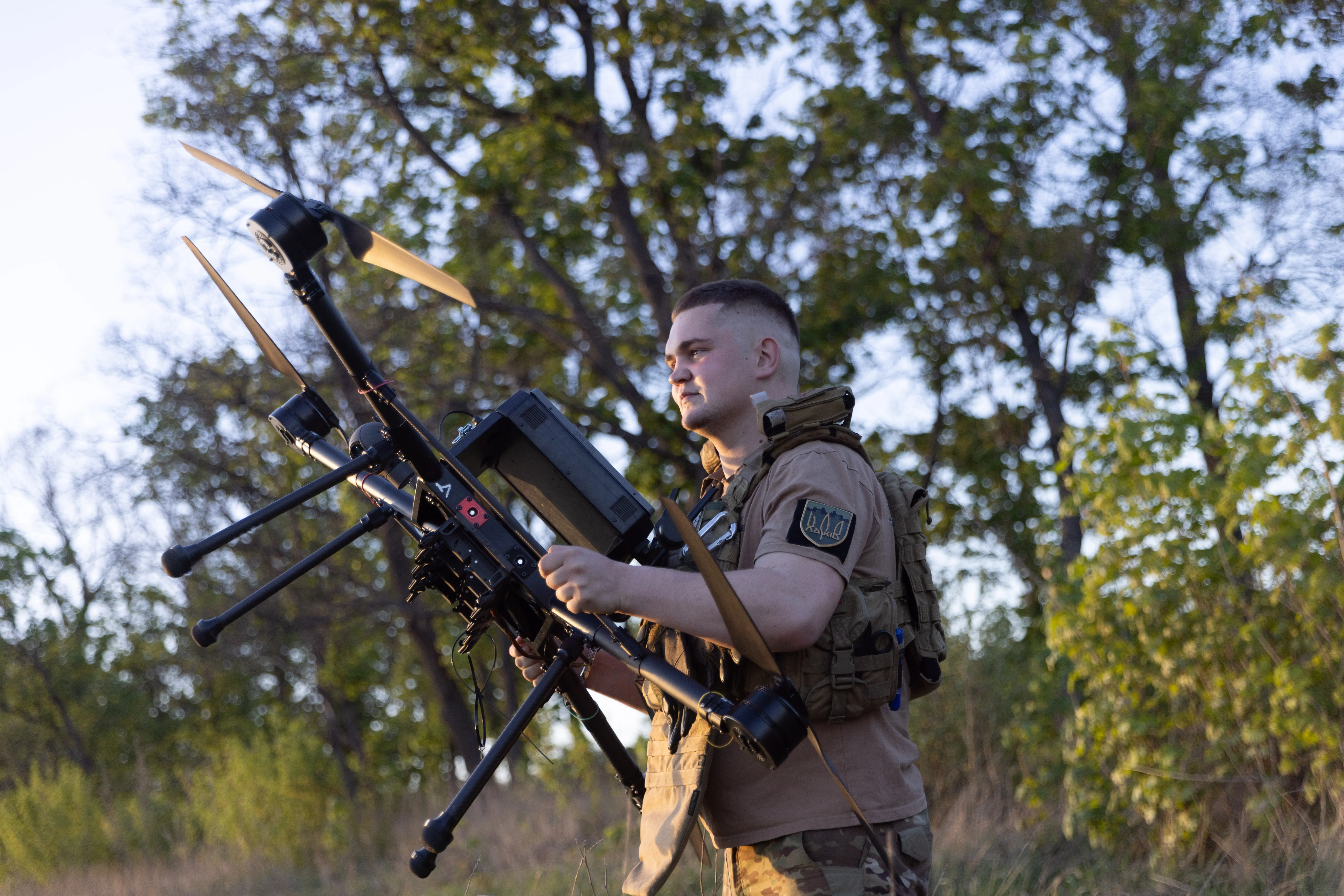 A man in khaki gear carries a large black drone among some trees