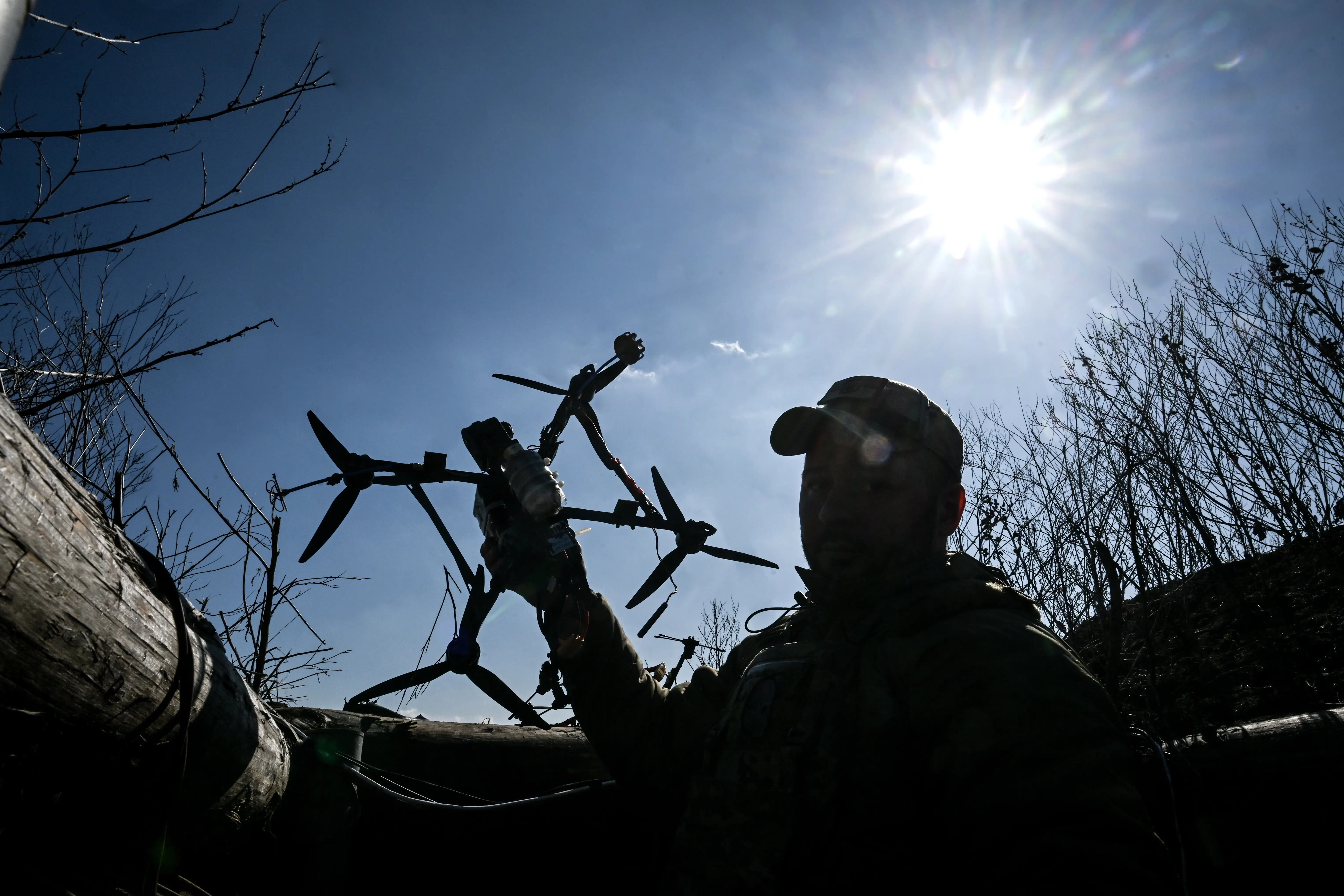 A man wearing a cap and holding a drone is sihouetted against the sun and a blue sky