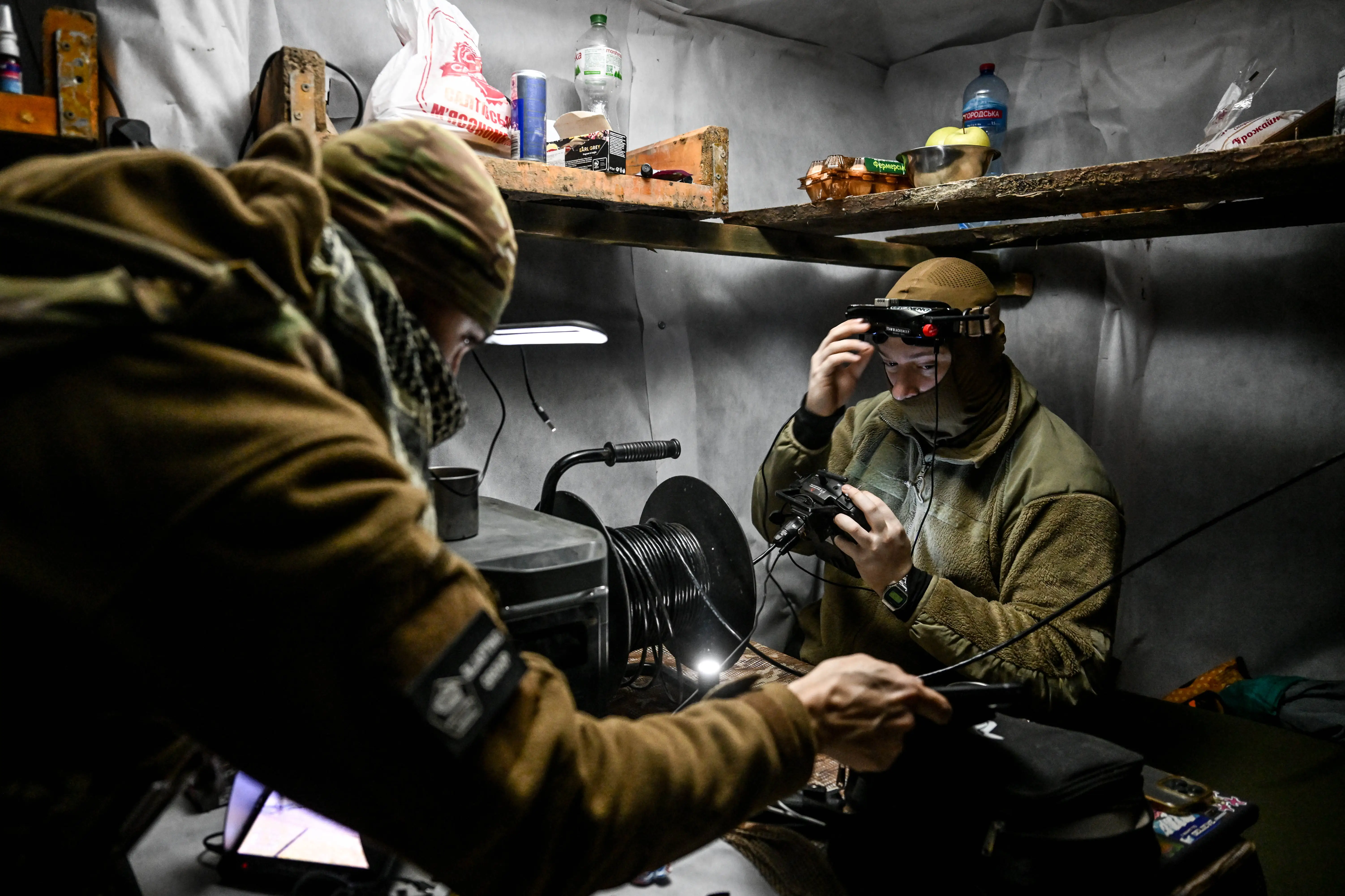 Two men in khaki gear and beanies in a small indoor location with a spool of cable, drone controllers, and water bottoles