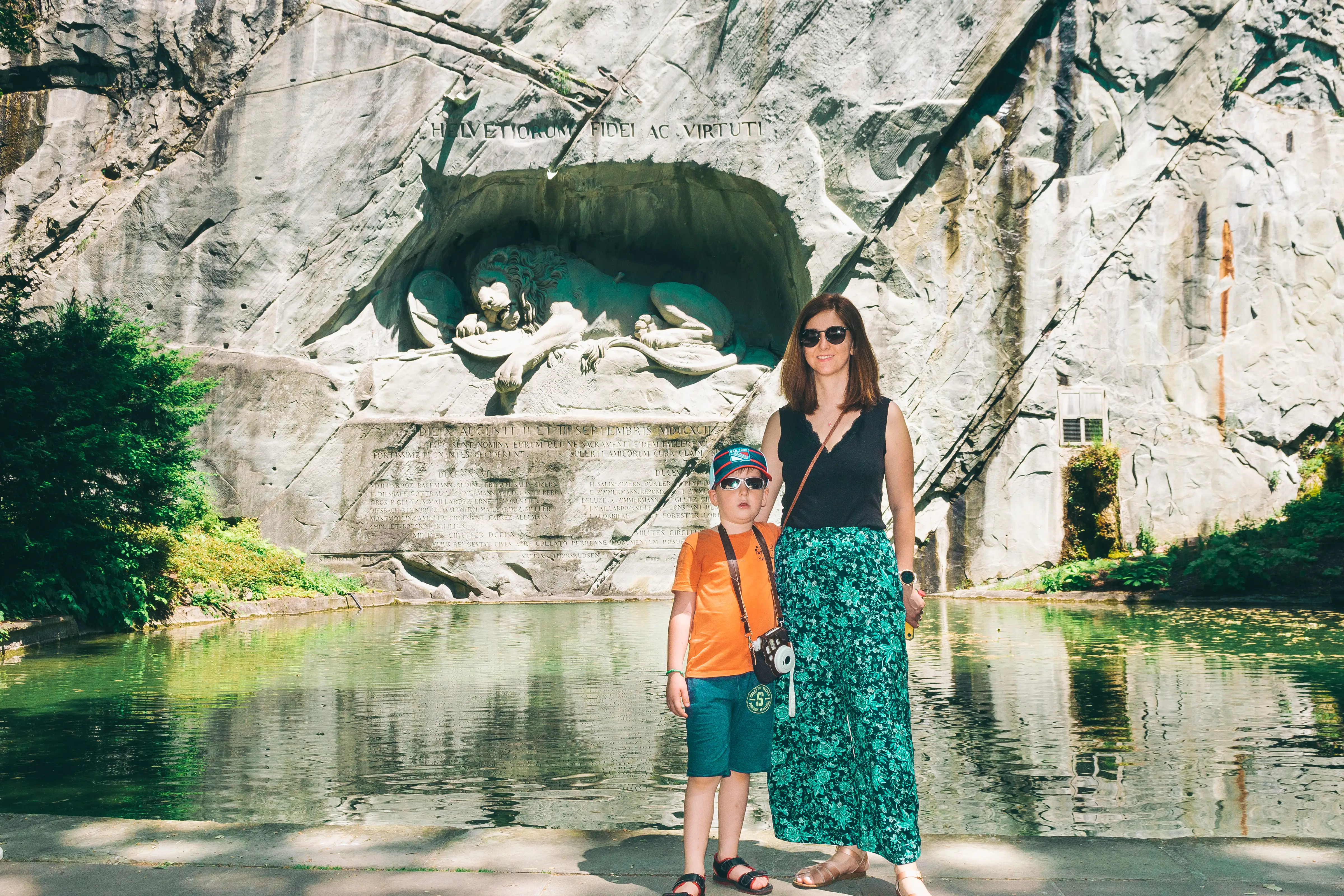 Woman smiling with boy, both wearing sunglasses, near body of water and stone structure in Switzerland