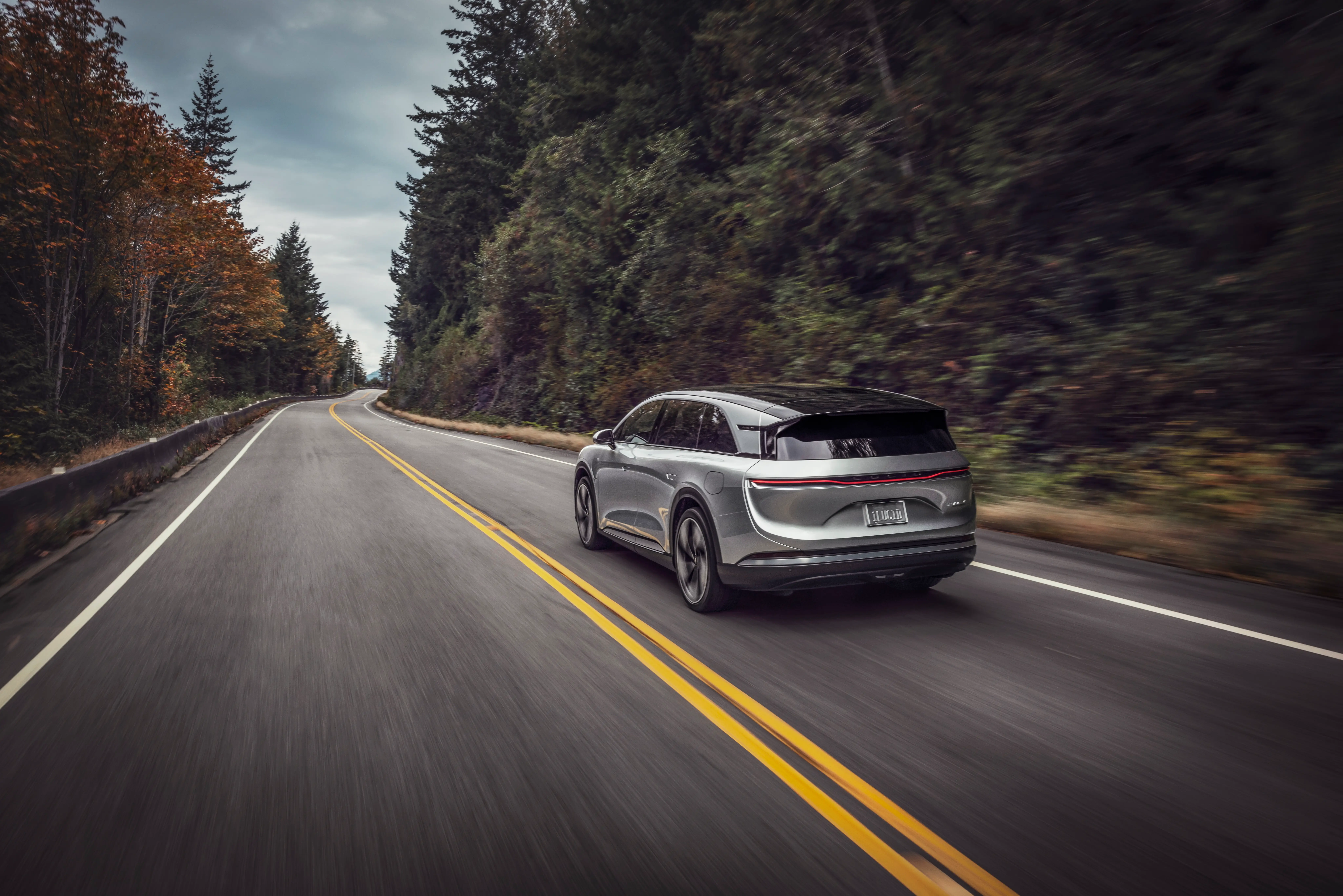 A Silver Lucid Gravity drives down a two-lane road past a wooded area.