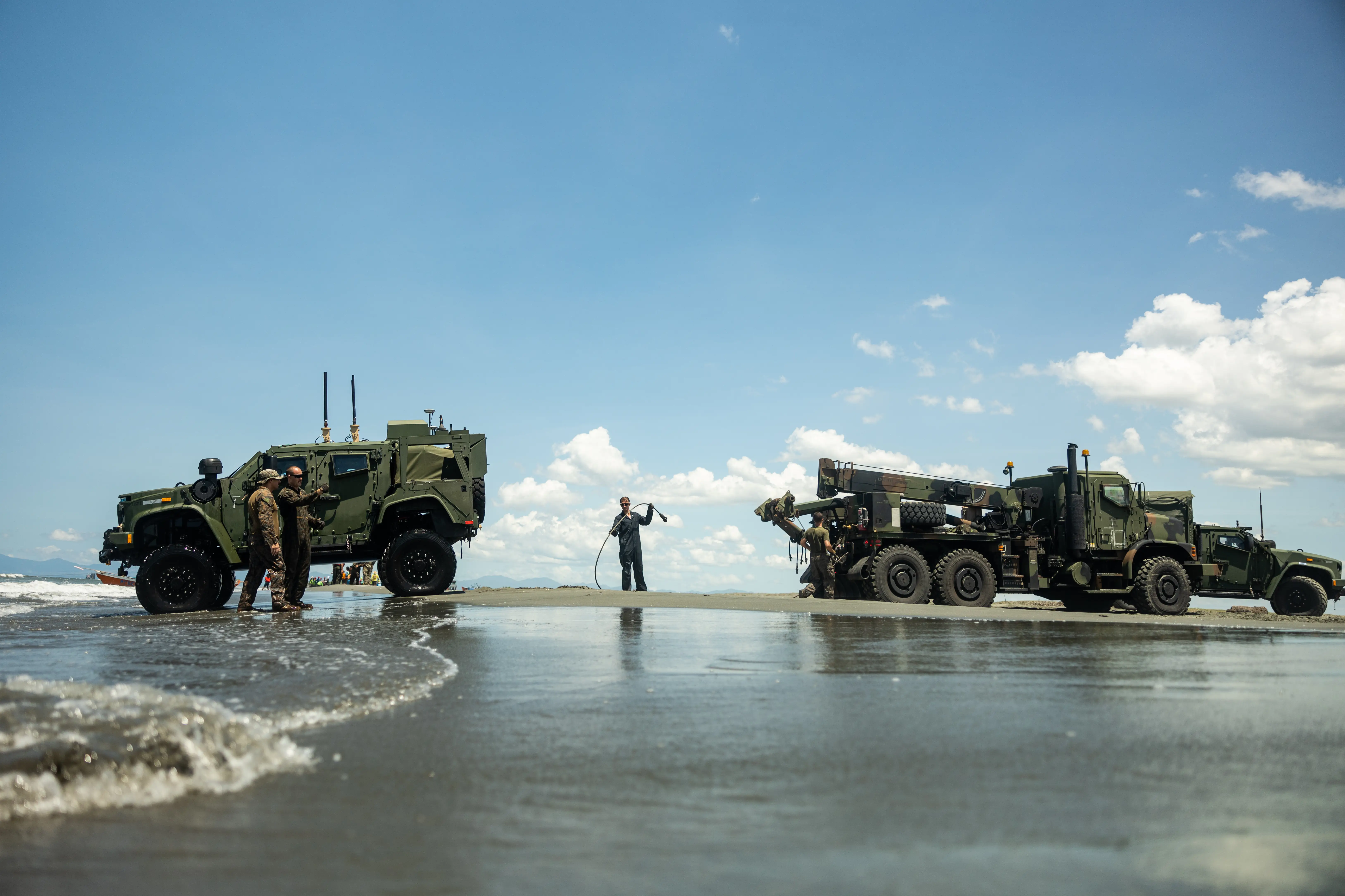 Two large vehicles sit on the beach with Marines standing around them. The sky is blue and cloudy.